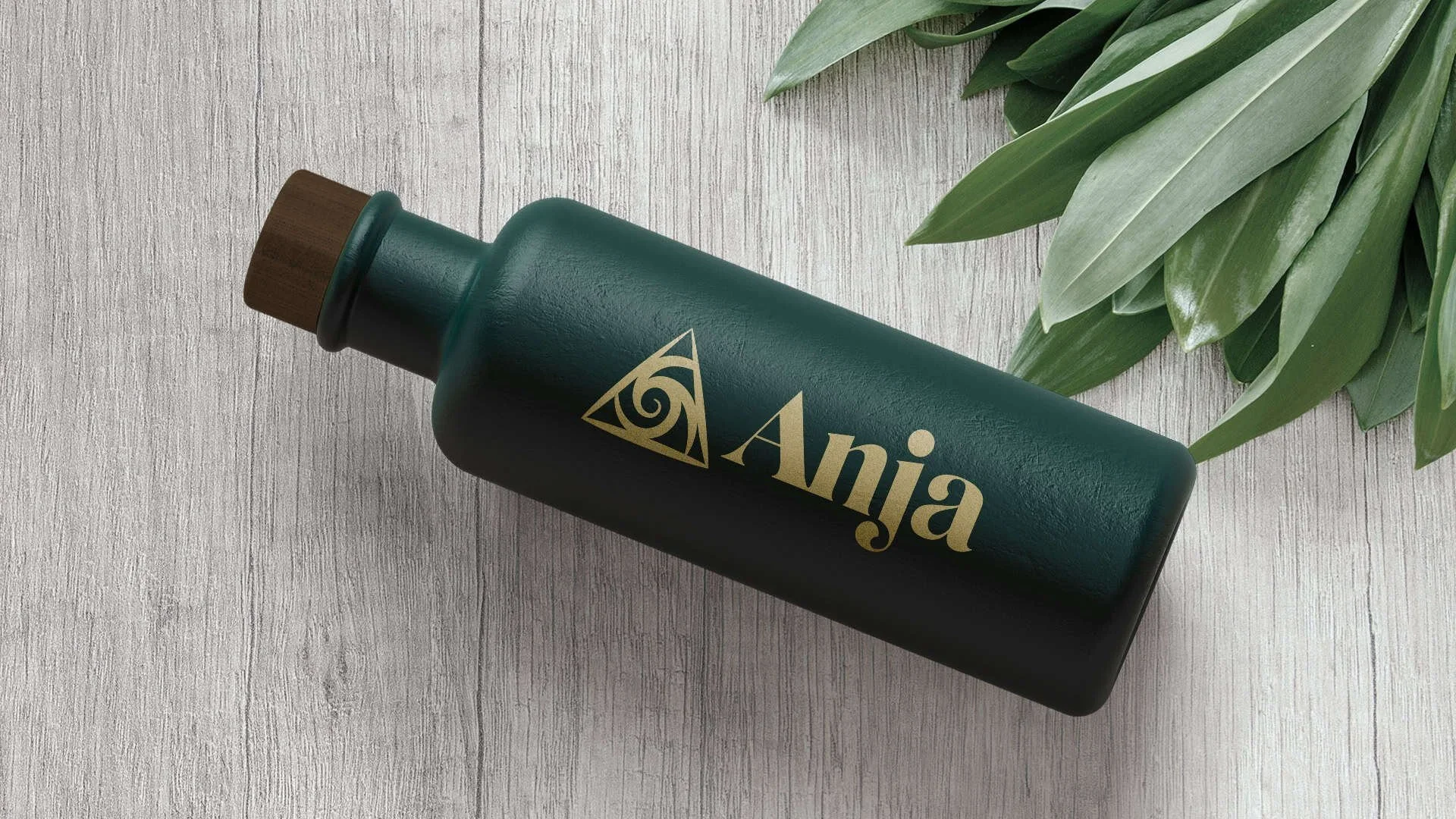 A dark green bottle with a brown cap and a gold logo and the word 'Anja' on it, lying on a light wooden surface next to green leafy plants.