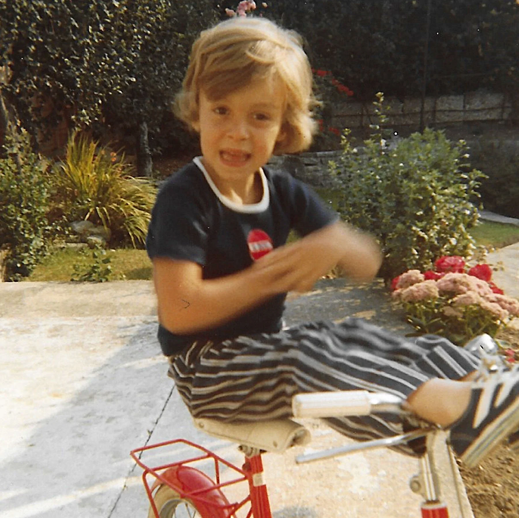Mark Nunez as a child riding a small red tricycle in a garden.