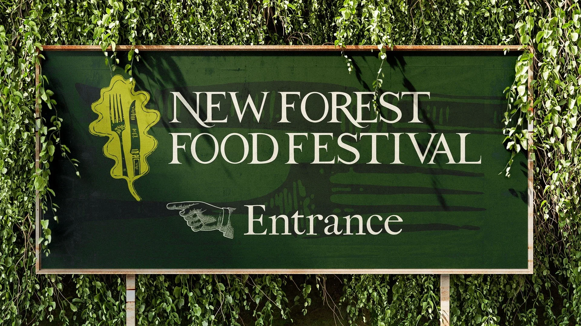 Green sign with white text reading "New Forest Food Festival Entrance," featuring a graphic of a fork and knife inside a yellow shape, mounted on two posts and surrounded by green foliage.