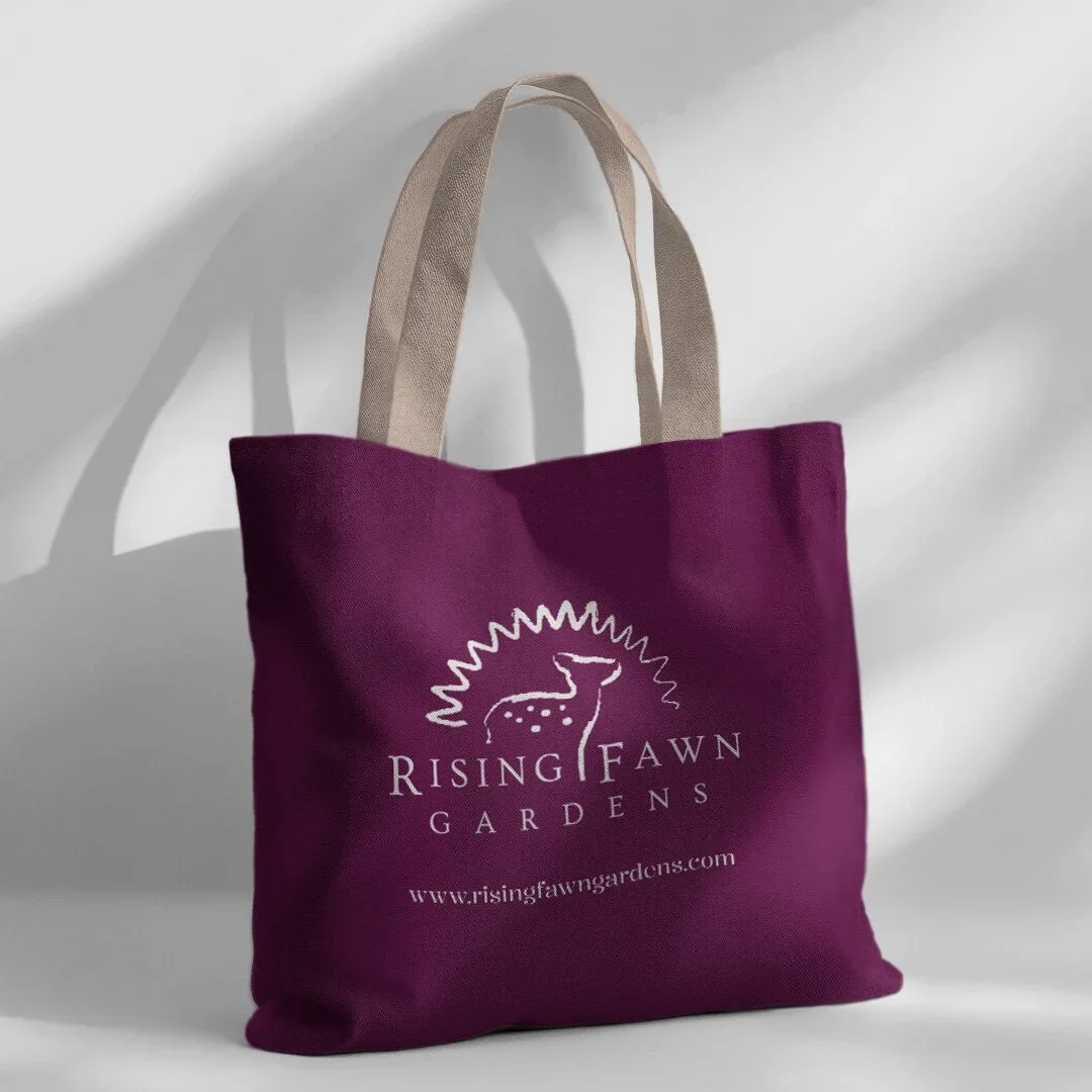 A purple tote bag with beige handles. The bag features a white logo of a deer with a stylized sun behind it and text that reads 'Rising Fawn Gardens' and the website address 'www.risingfawngardens.com.'