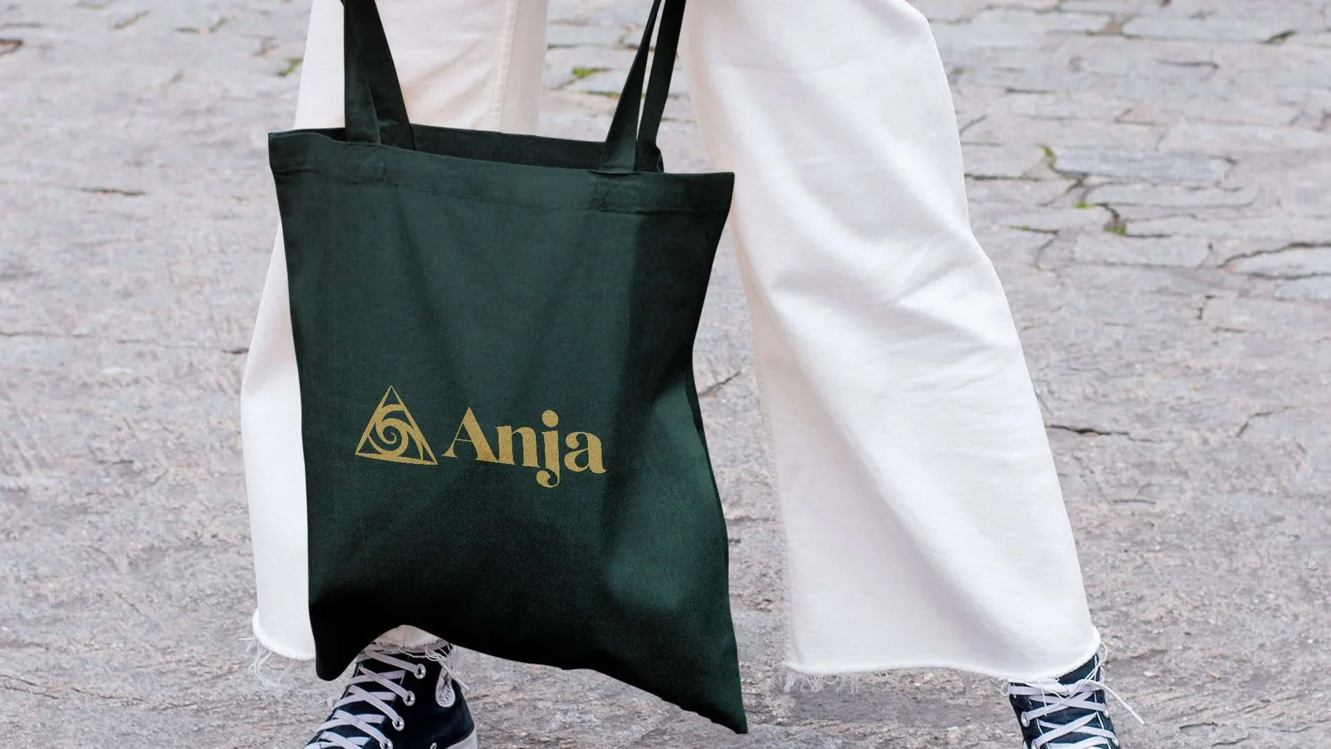 Person holding a green tote bag with the word 'Anja' and a spiral triangle logo printed in gold, standing on a stone pavement, wearing white pants and black and white sneakers.
