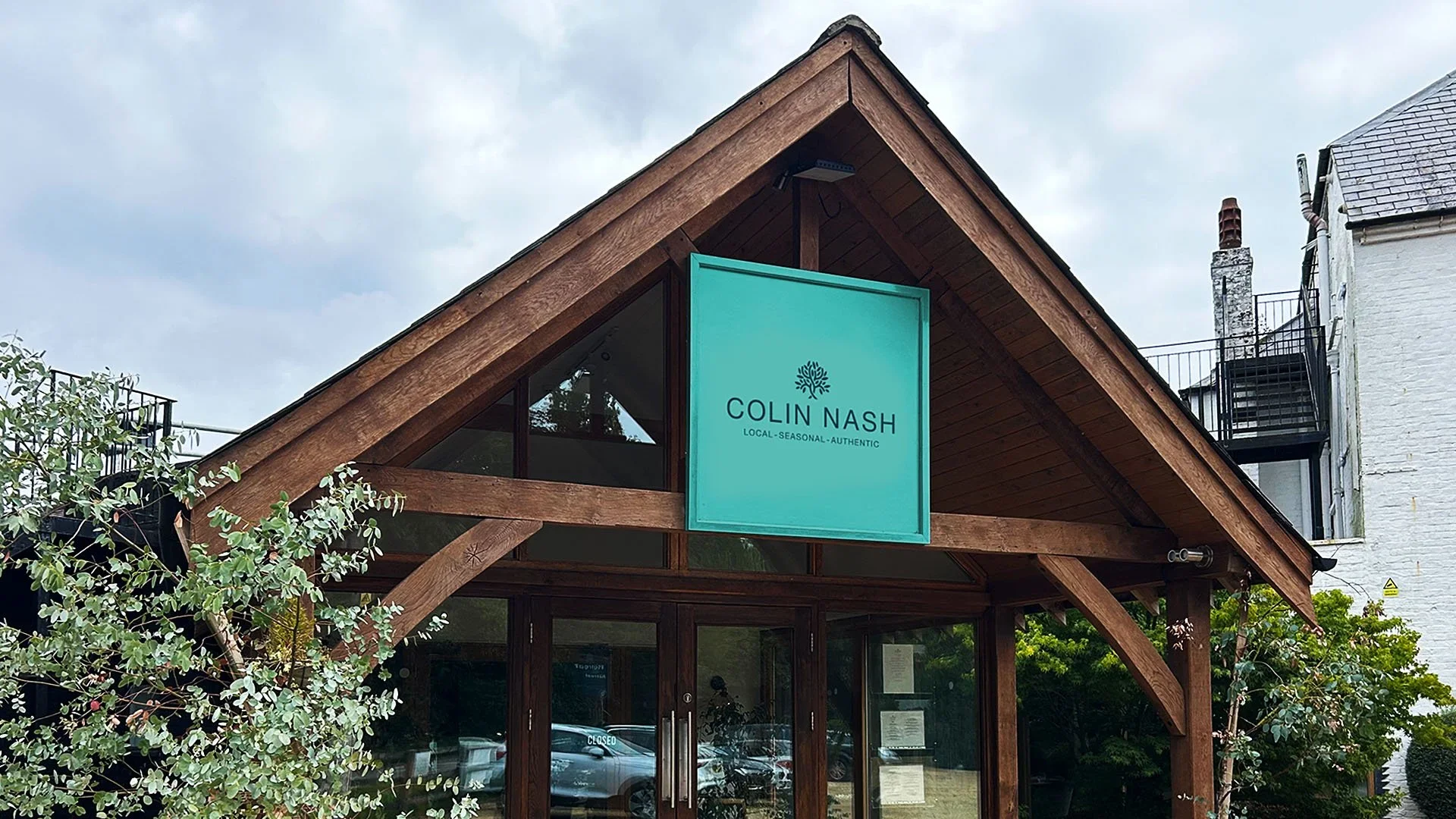 Exterior view of a building with a prominent turquoise sign displaying 'Colin Nash, Local-Seasonal-Authentic' above the entrance. The building has a wooden triangular facade and glass doors. There are trees and parked cars nearby.
