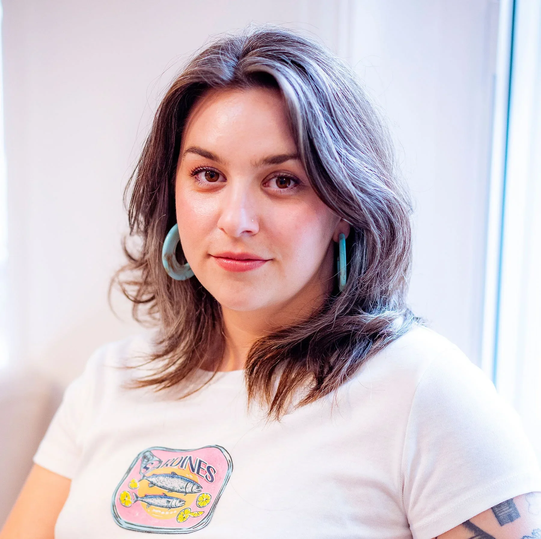 Delila Miske wearing a white top and turquoise earrings in a natural light portrait.