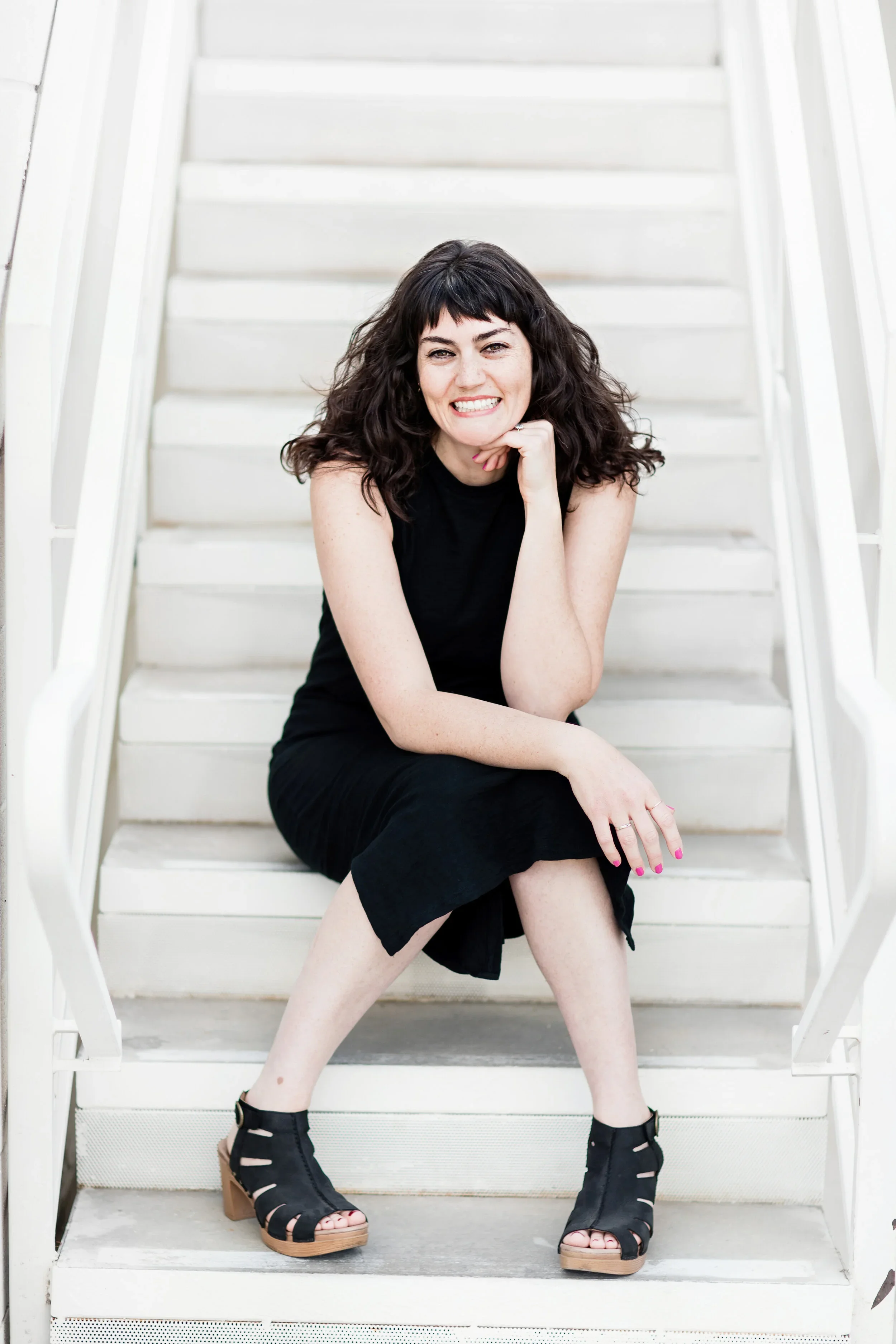 A woman with curly dark hair, wearing a black sleeveless top, black skirt, and black open-toe heels, sitting on white stairs and smiling at the camera.