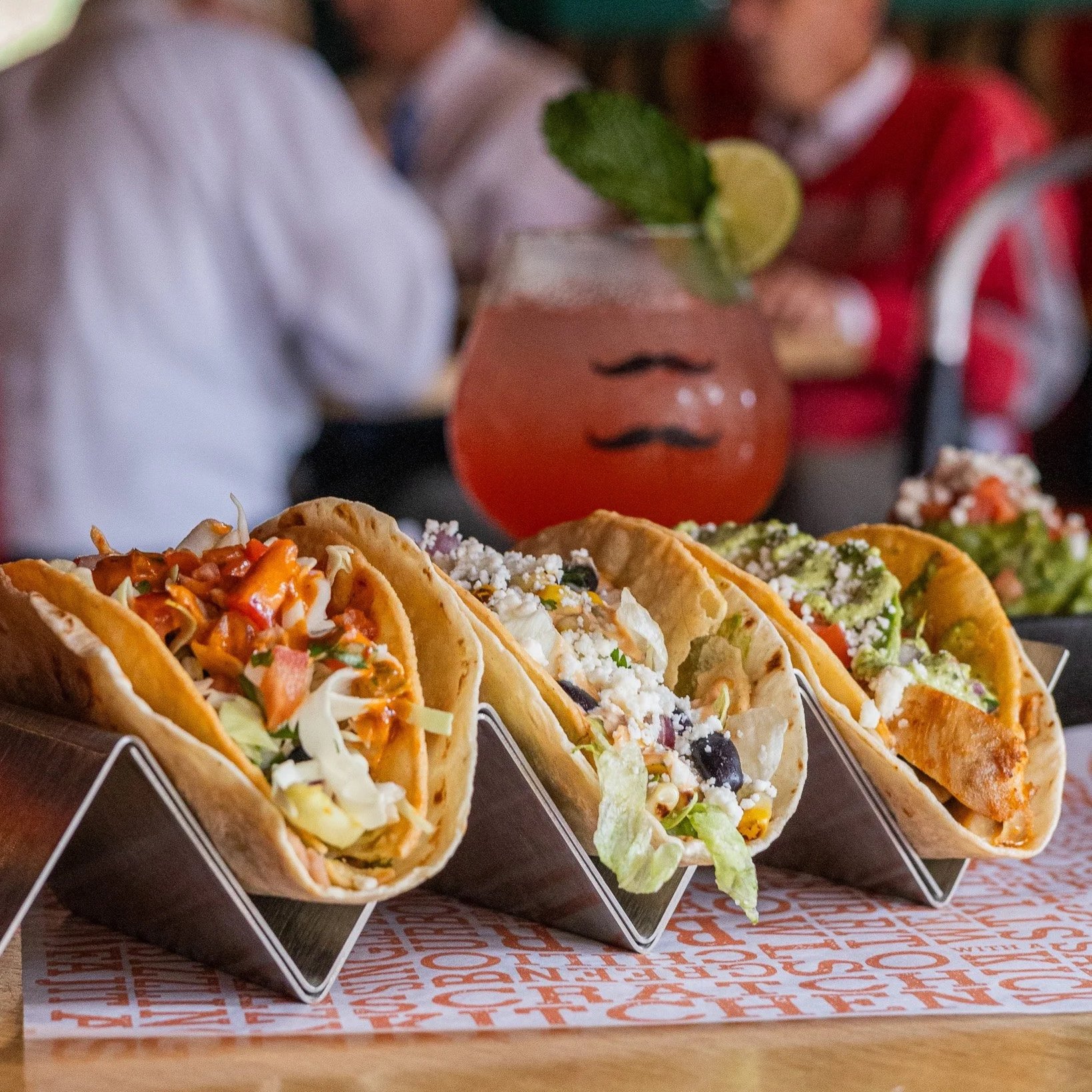 Three tacos in metal holders topped with fresh ingredients, served on a table with a pink cocktail in the background at Orange Umbrella Restaurants