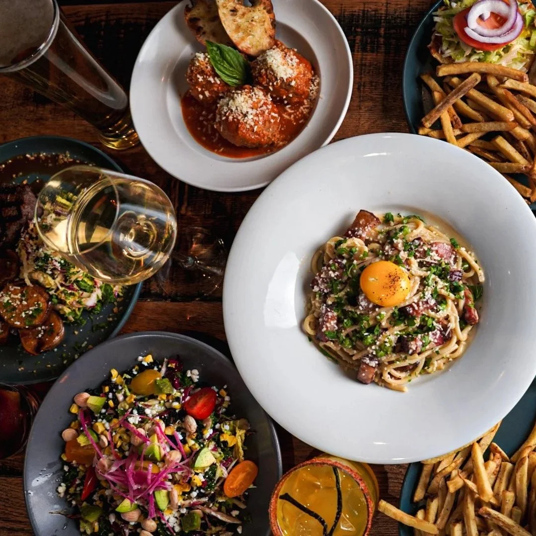 An overhead view of plated restaurant dishes including pasta with egg yolk, meatballs with bread, a colorful chopped salad, fries, and glasses of wine on a wooden table.