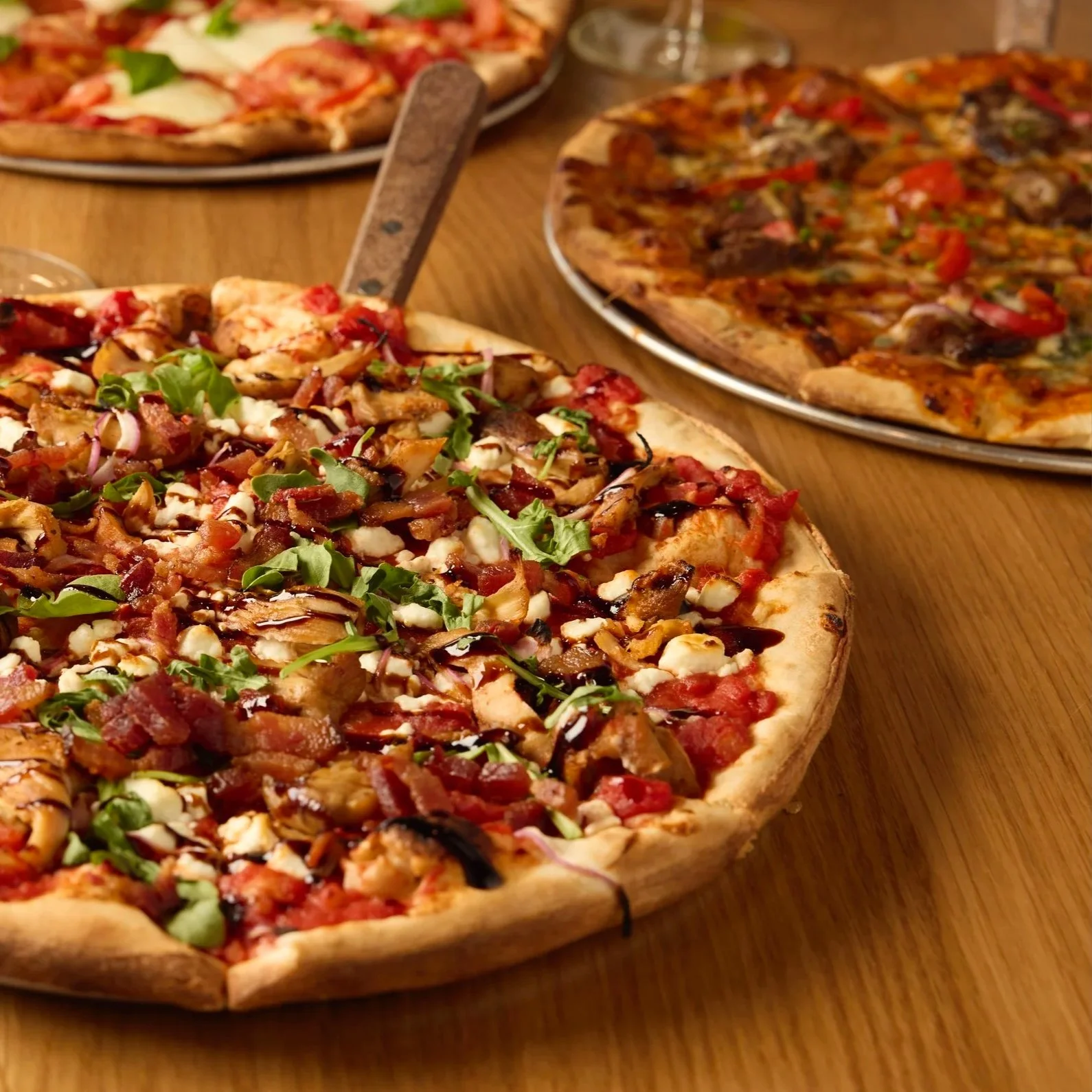 Assorted wood-fired pizzas topped with fresh mozzarella and basil on a dining table, surrounded by wine and cocktails.