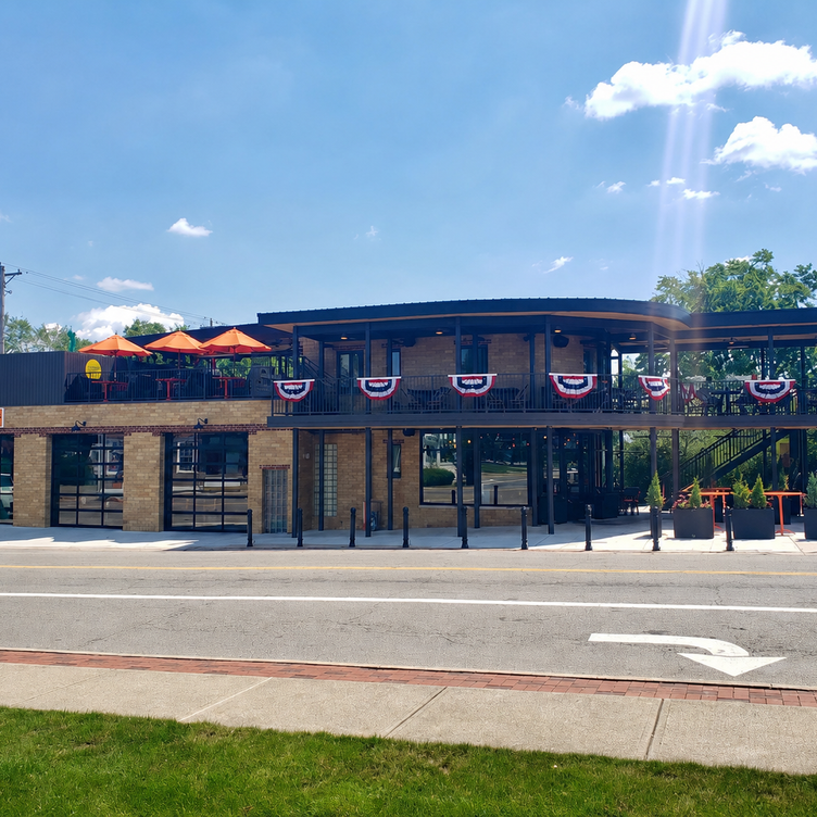 A two-story brick building decorated with red, white, and blue bunting, with an outdoor terrace on the upper level featuring orange umbrellas and seating, on a bright sunny day with a blue sky and scattered clouds.