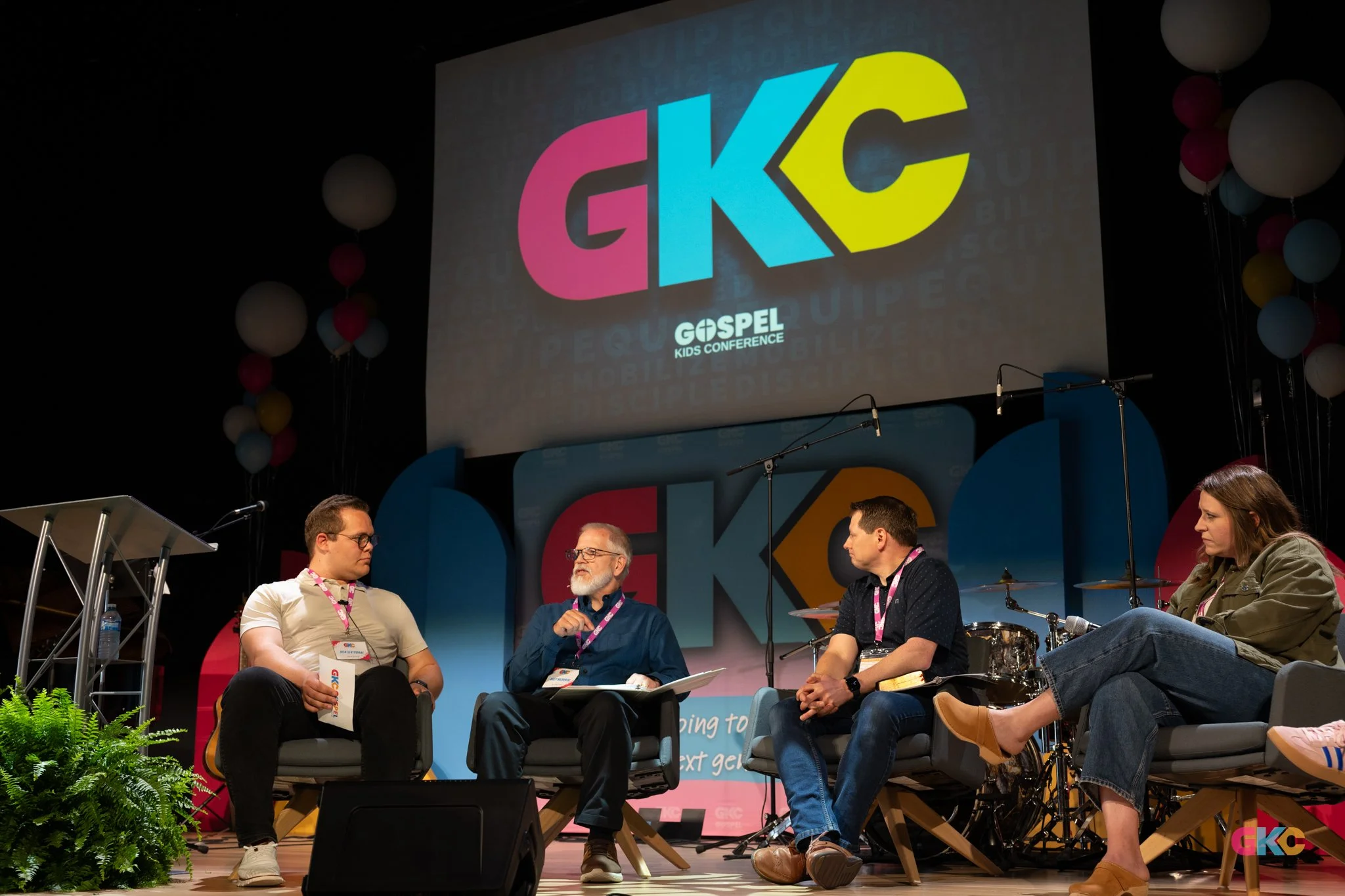 Panel discussion at a gospel kids conference, with four people seated on stage, large GKC logo on a screen behind them, microphones, drums, balloons, and a colorful background.