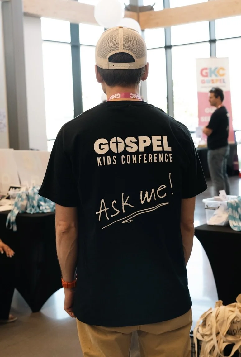 Back of a person wearing a black T-shirt with the text 'GOSPEL KIDS CONFERENCE' and 'Ask me!' written on it, inside a well-lit room with large windows, tables, and other people in the background.