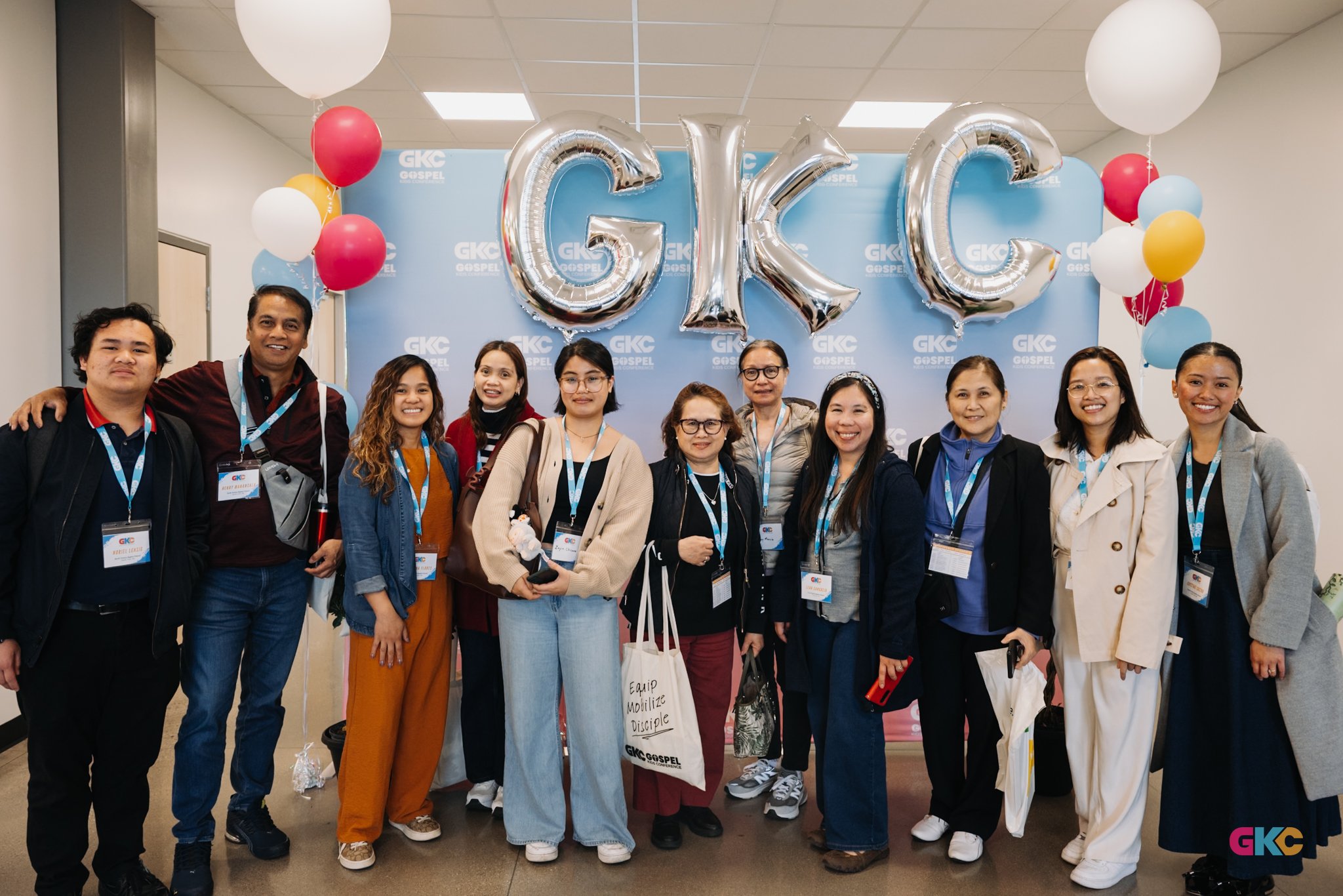 Group of people standing together at an event, smiling, with balloons and a blue backdrop that has large silver balloons spelling 'GKC' and multiple smaller 'GKC Gospel' logos.