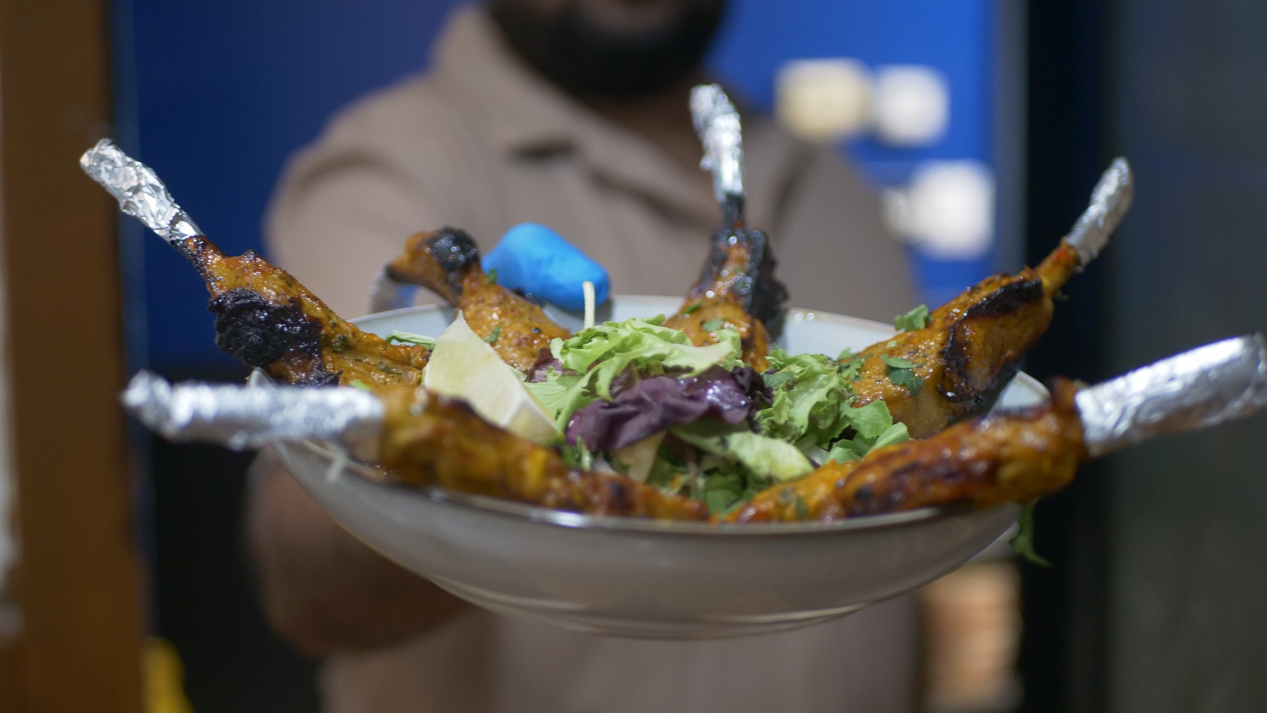 A person holding a plate of grilled chicken drumsticks wrapped in foil, garnished with salad and lemon slices.