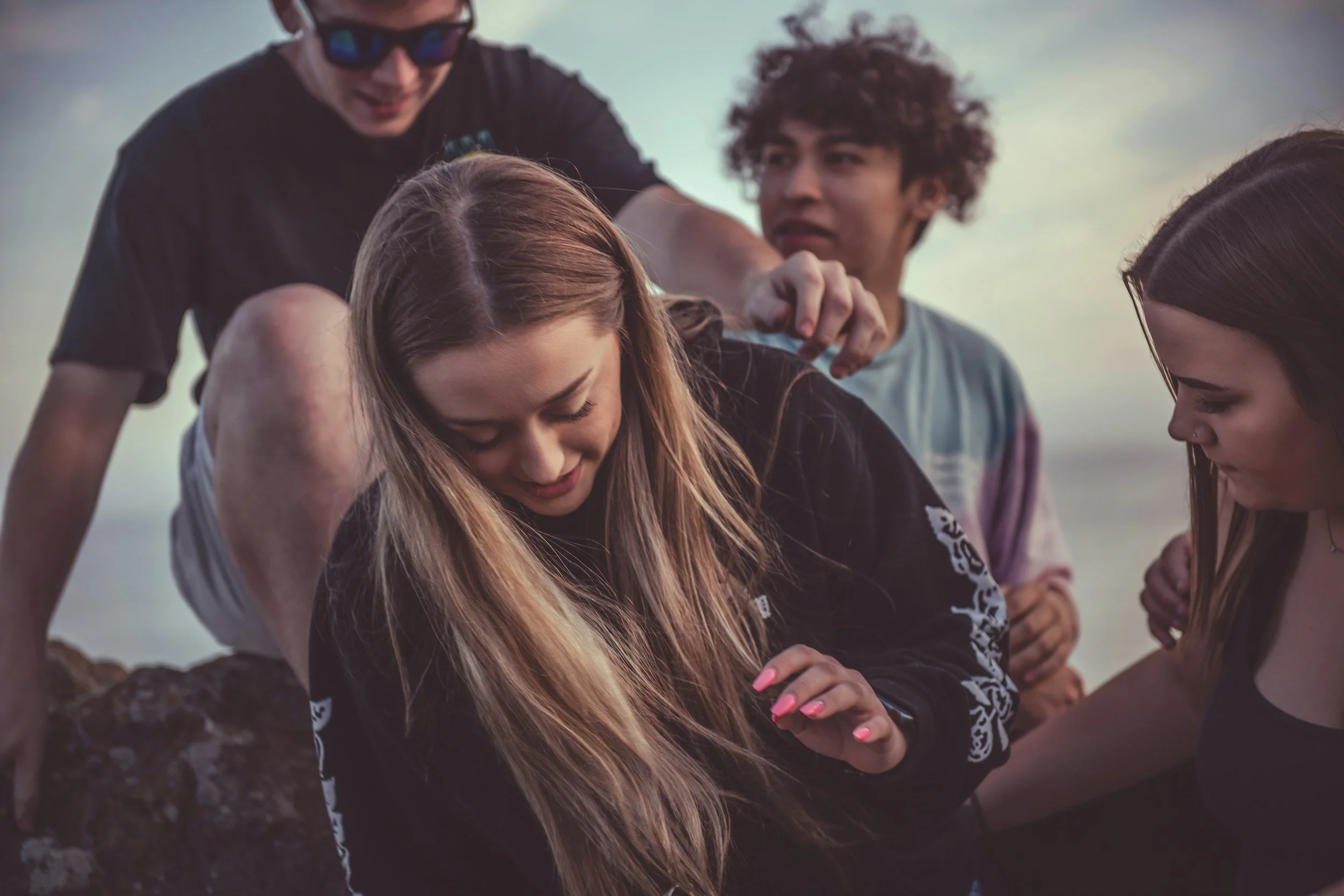 A group of four young people gathers outdoors near rocks, with the sky in the background. They are closely interacting, with one girl in the group smiling and looking down, and others observing her attentively.