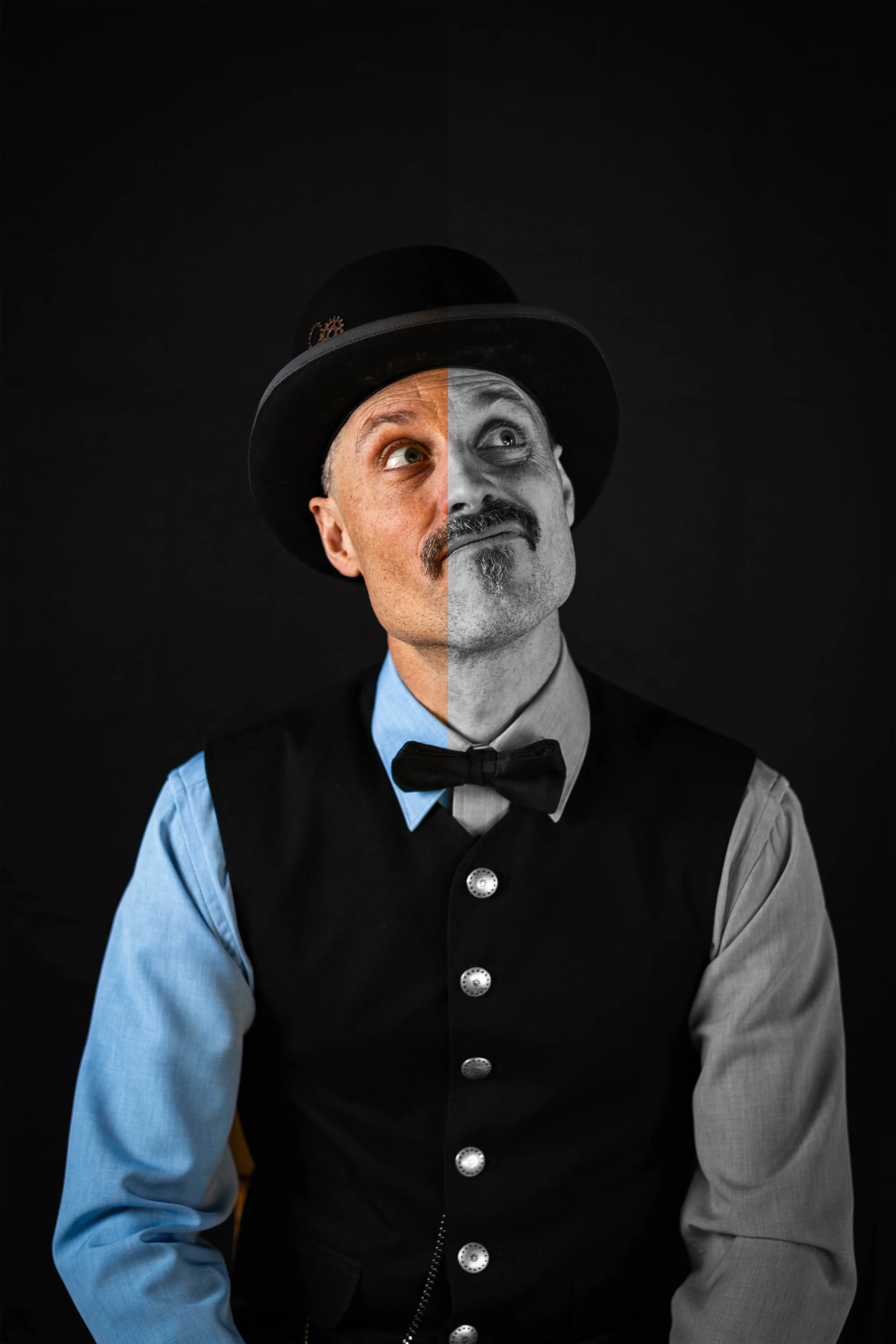 A man dressed as a Victorian-era circus ringmaster, split between color and black-and-white, wearing a top hat, bow tie, and vest, looking upward with a playful expression, against a dark background.