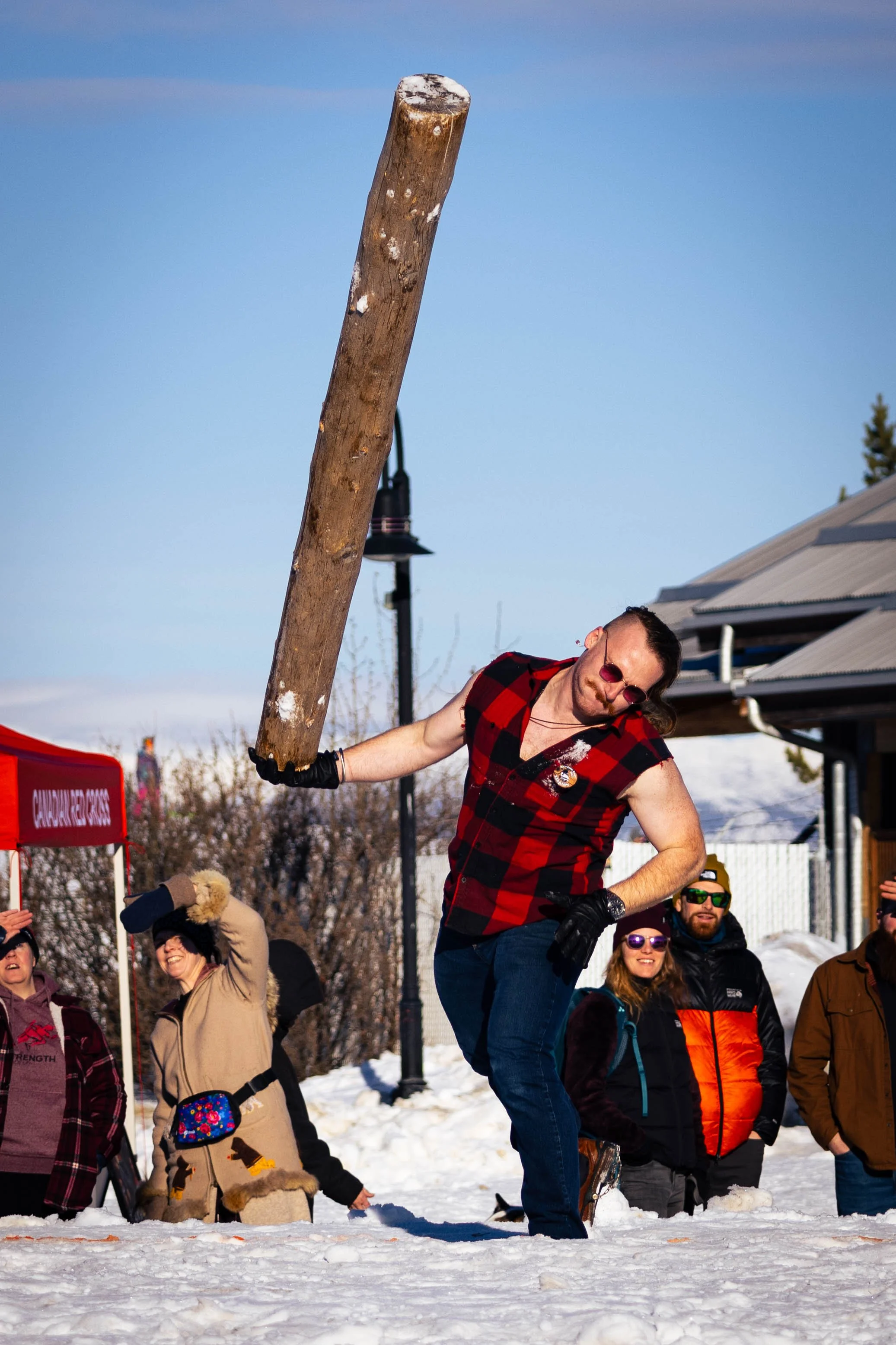 A man with sunglasses wears a red and black flannel shirt and black gloves, participating in a snow hockey game while holding a large wooden club or bat.