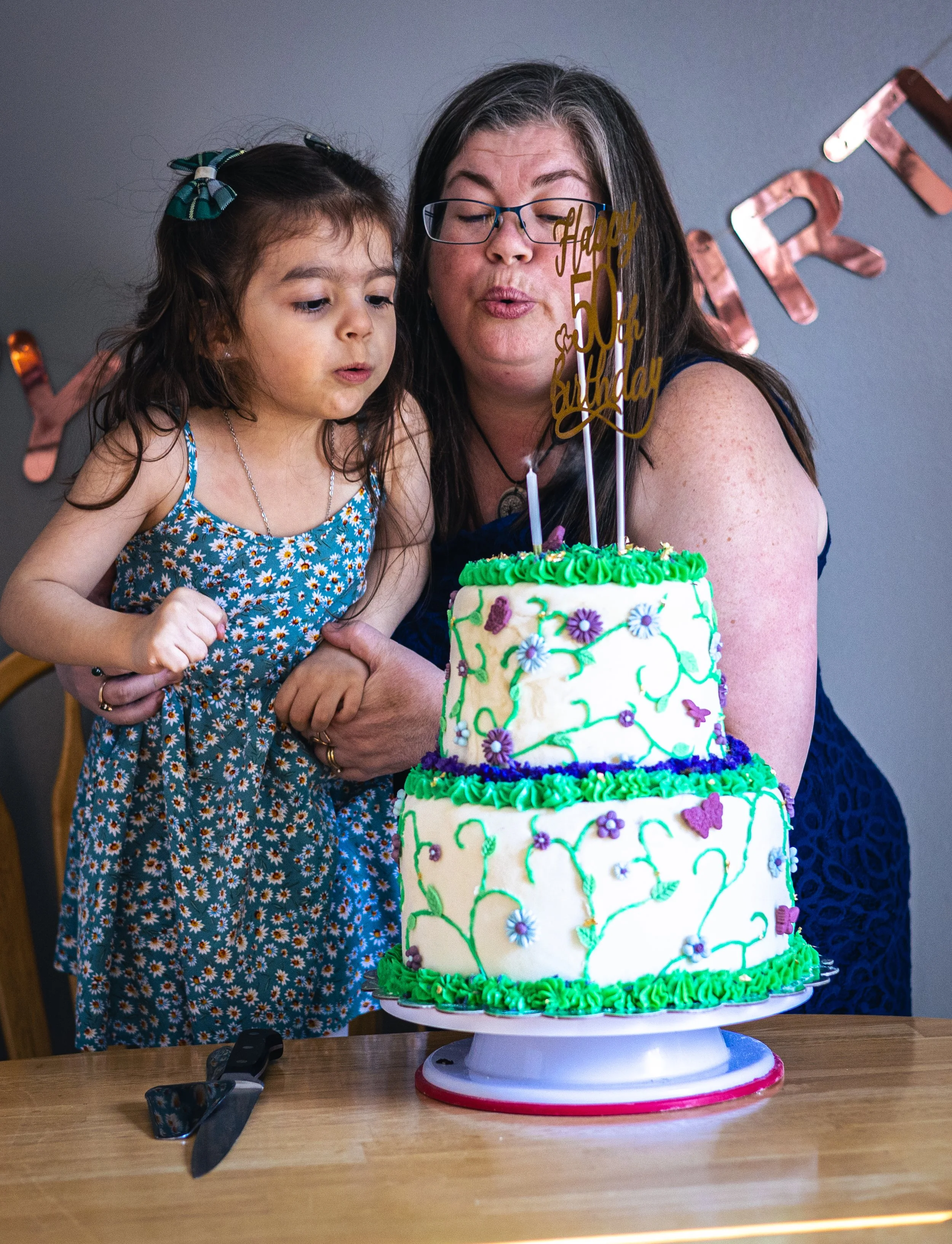 A woman and a young girl blow out candles on a two-tiered birthday cake decorated with green and purple icing, flowers, and a 'Happy 50th Birthday' topper.