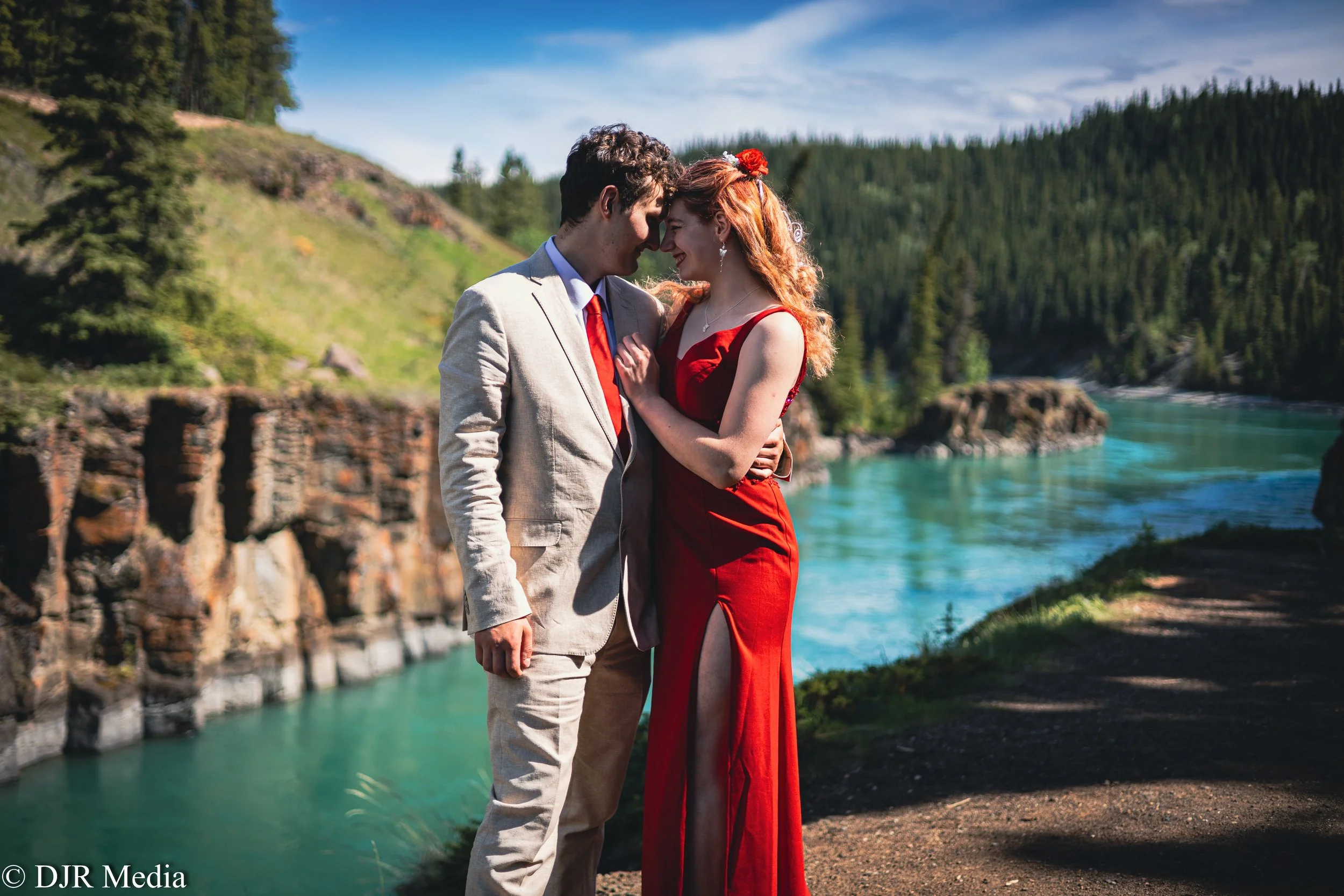 A couple stands close together by a river in a scenic outdoor setting, embracing and smiling at each other, with trees and hills in the background.