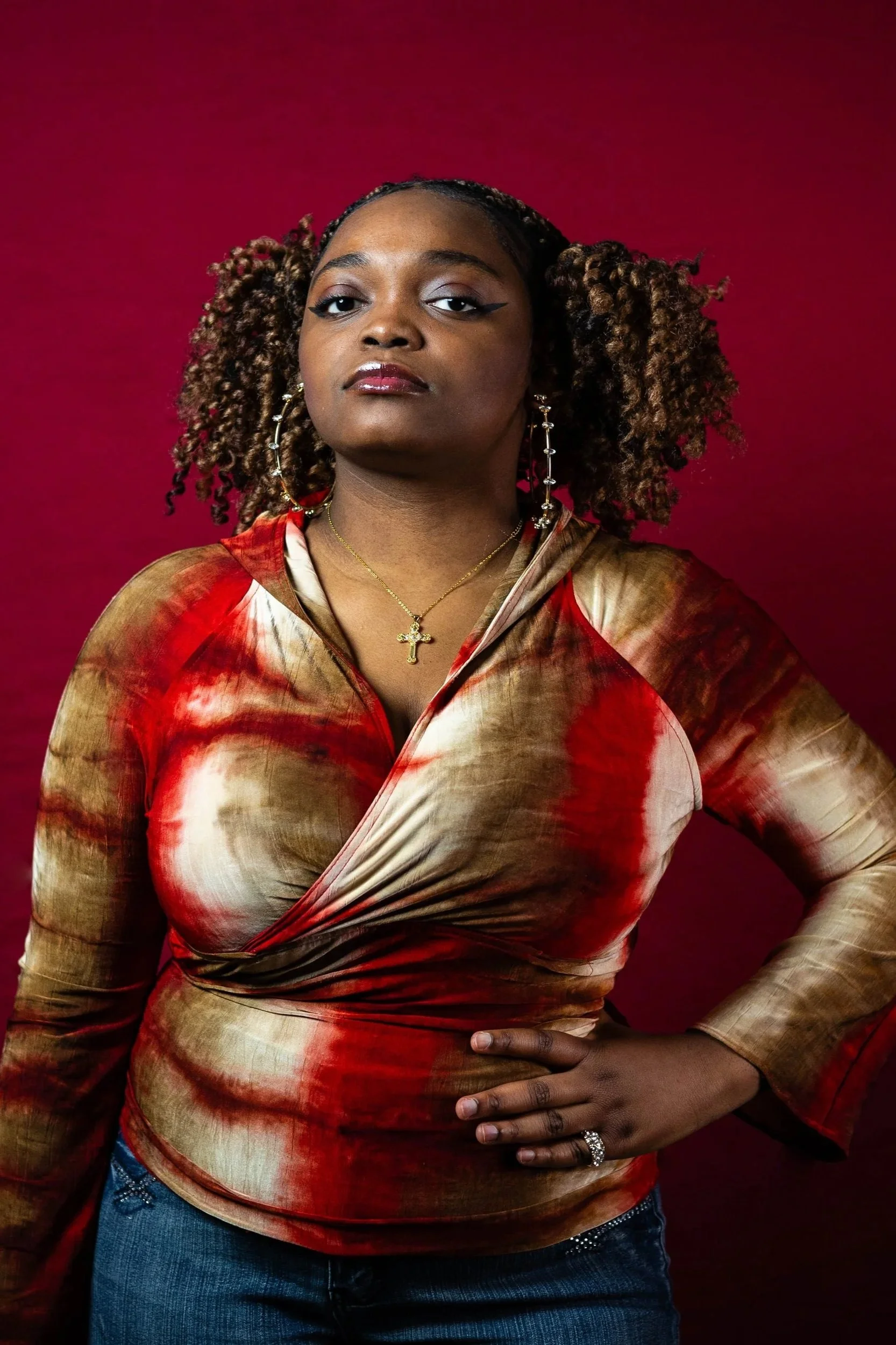 Portrait of a confident woman with curly hair wearing a red and beige tie-dye long-sleeve top, jeans, and jewelry including rings, earrings, a necklace with a cross pendant, and makeup with winged eyeliner, posing with one hand on her hip against a red background.