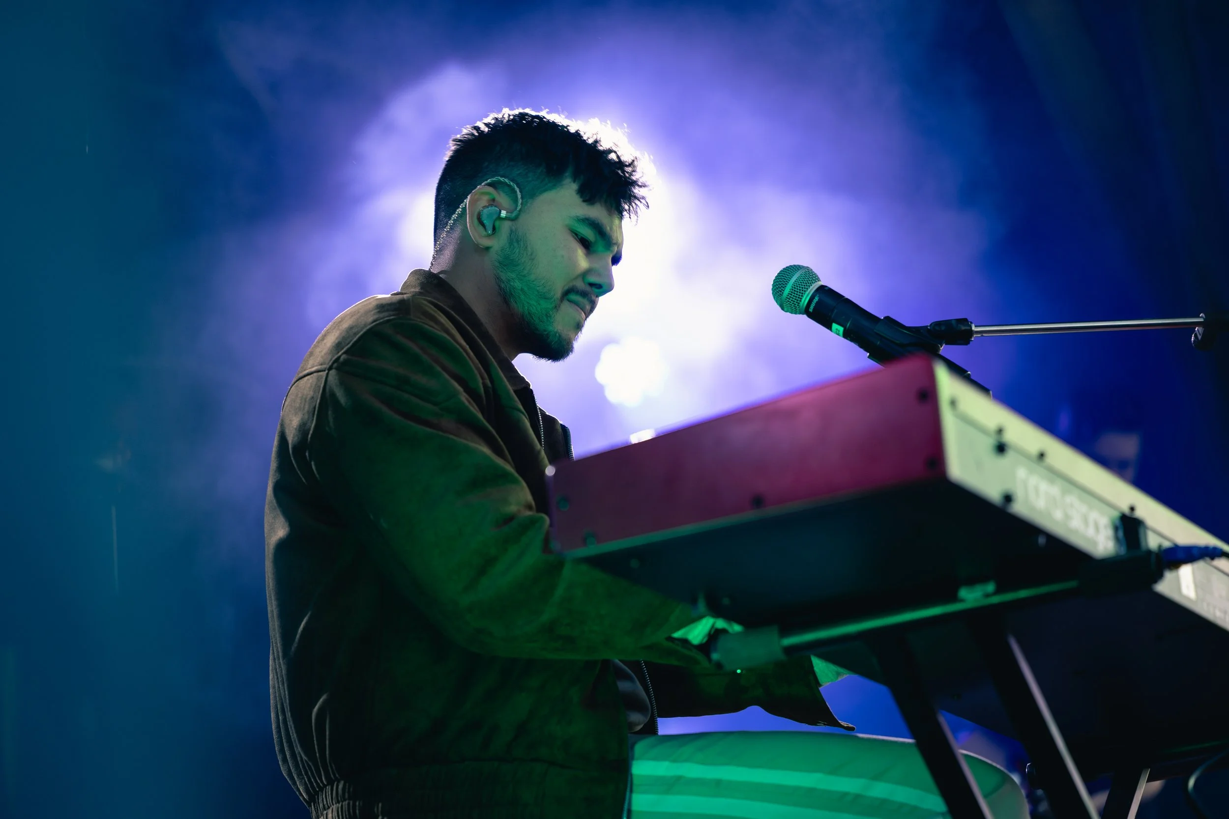 A young man playing keyboard on stage, wearing a brown jacket and earpiece, with purple and blue lighting in the background.