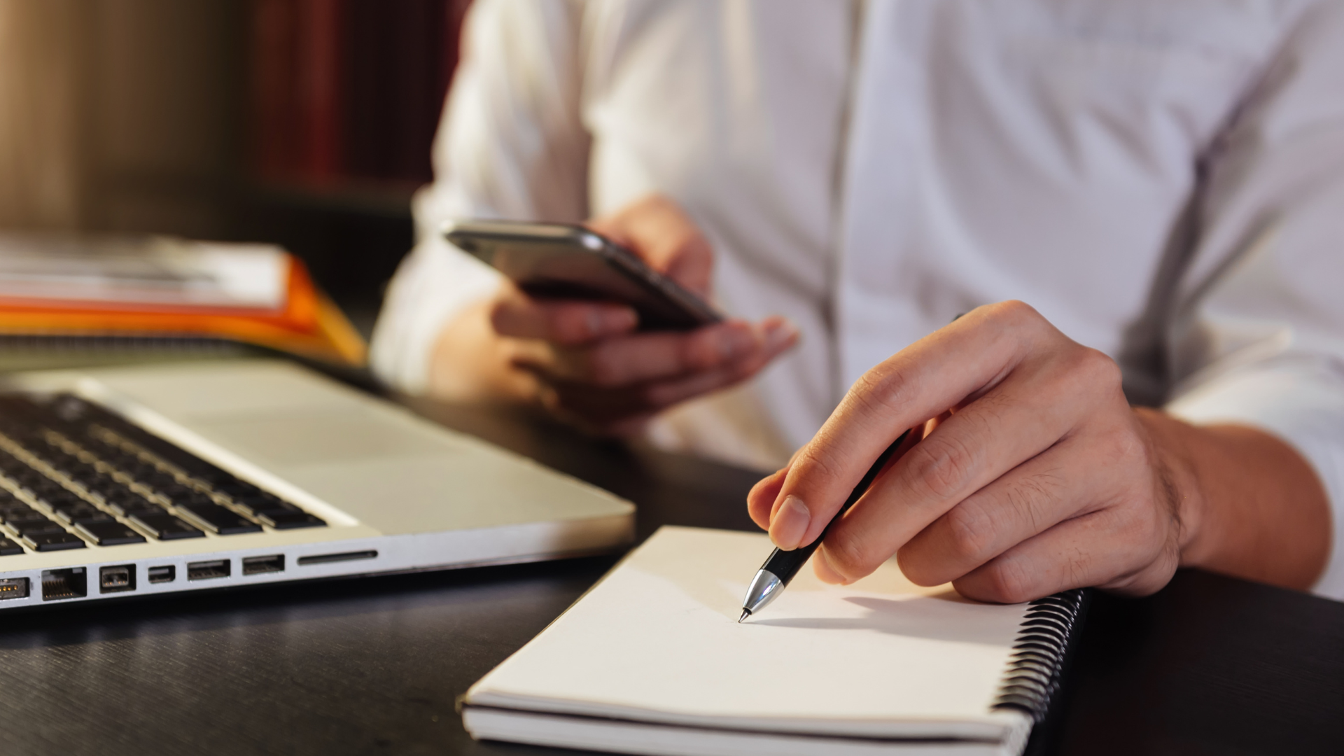 Close-up of business owner's hands writing profit and compensation calculations in a notebook beside an open laptop