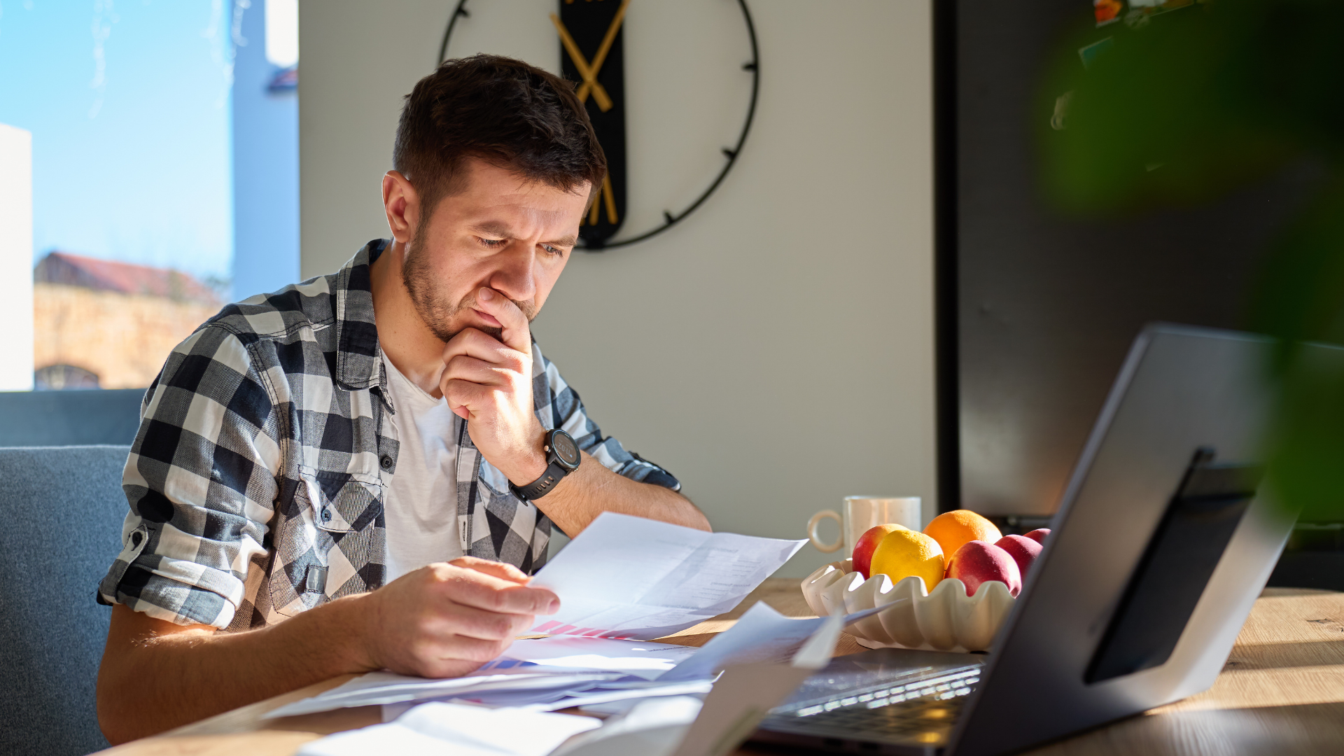 Small business owner sitting at a wooden desk reviewing financial documents on a laptop in a bright home office