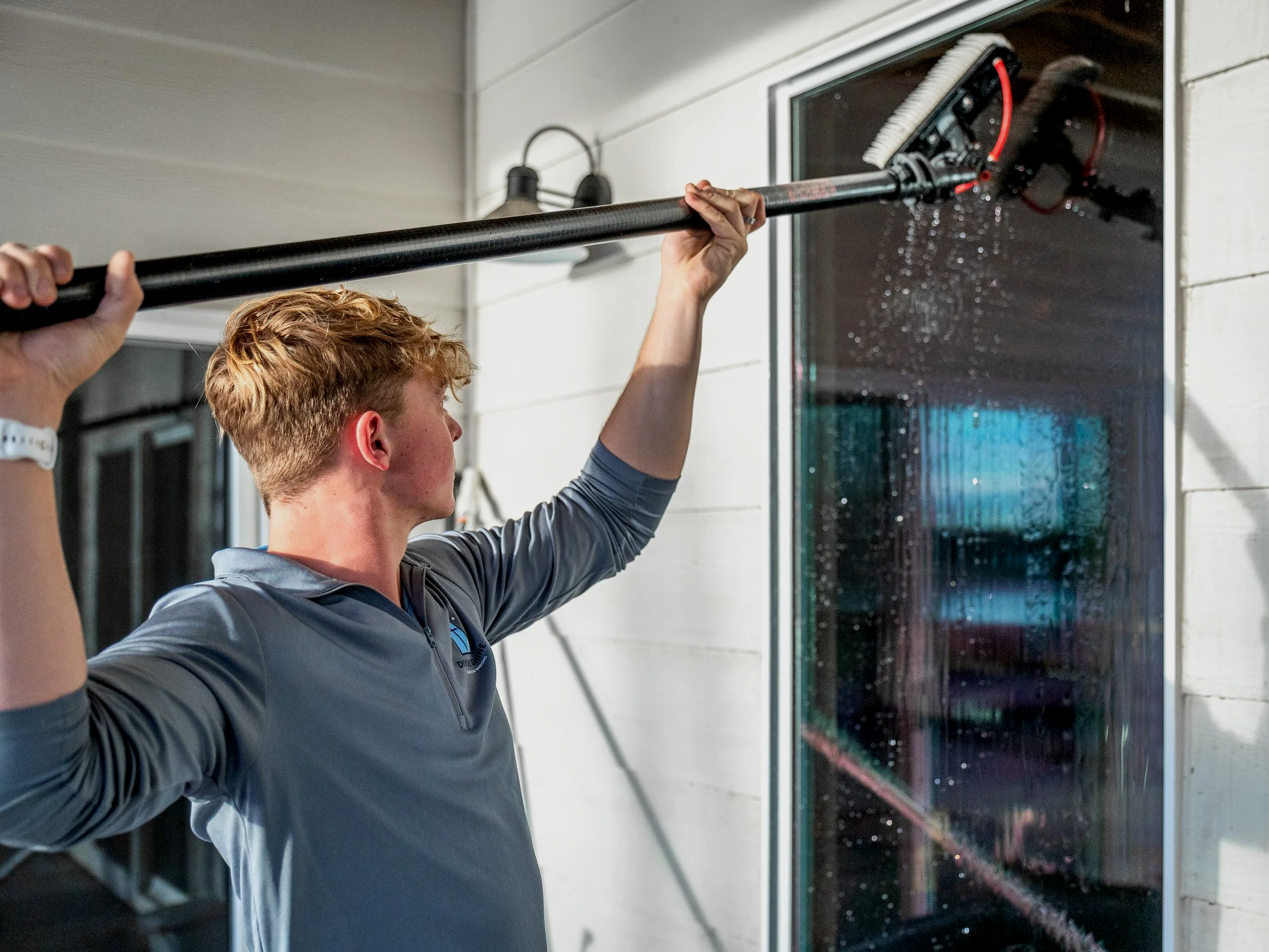 Young man cleaning a window with a long-handled scrub brush inside a building.