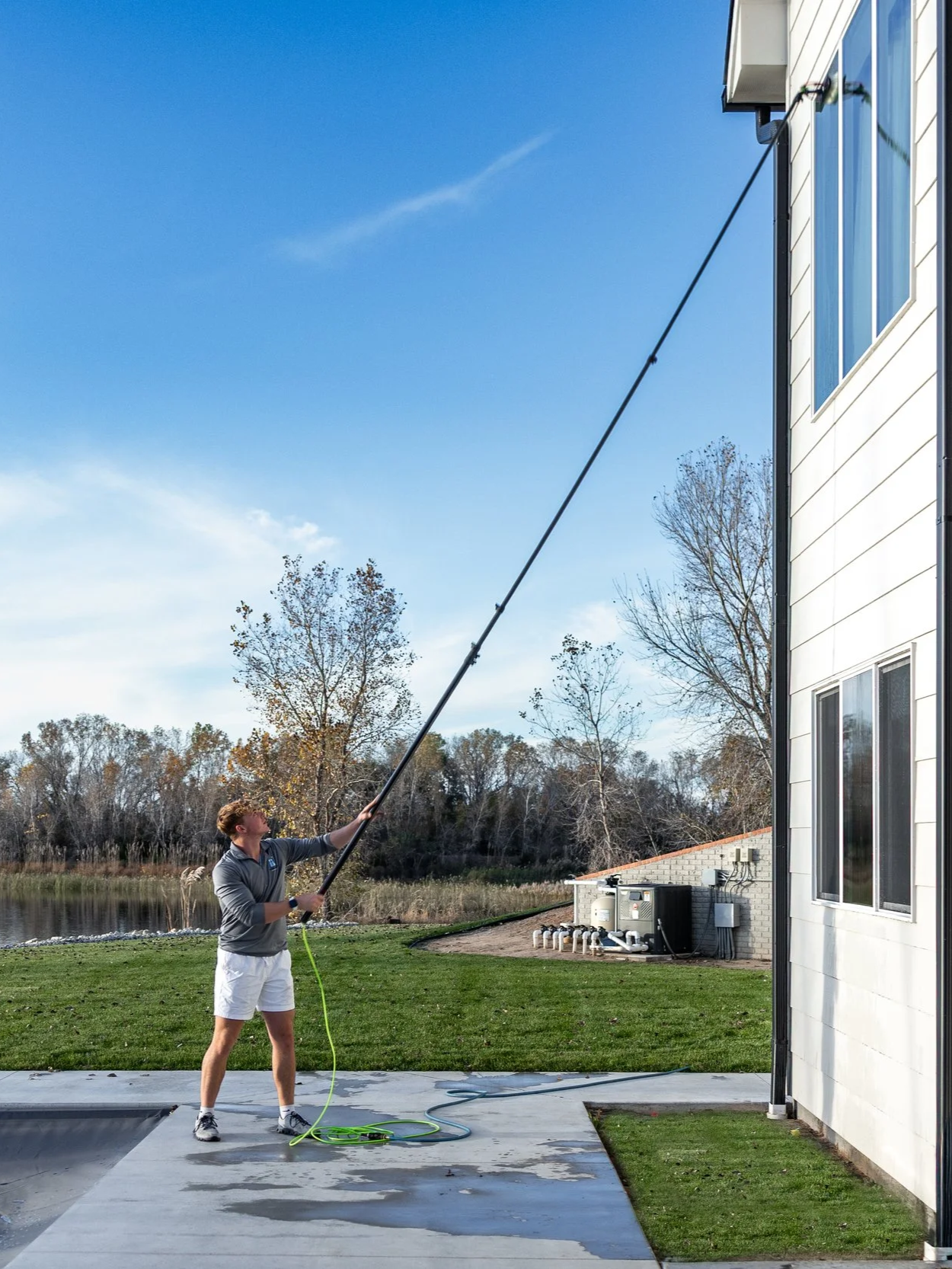 A person is cleaning windows on the side of a white house using an extension pole, with a pond and trees in the background under a blue sky.