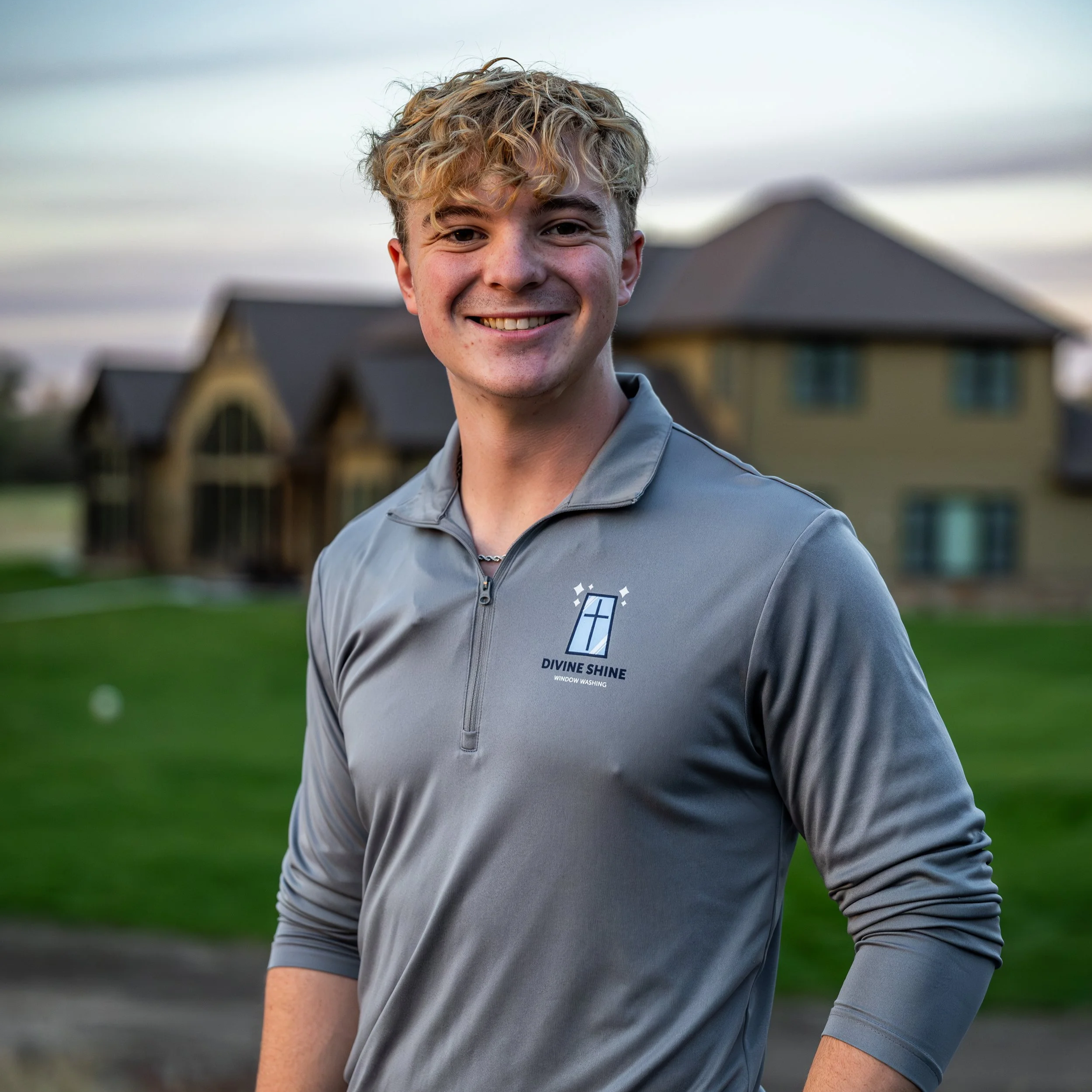 Young man with curly blonde hair smiling outdoors at sunset, wearing a gray quarter-zip shirt with a logo that reads 'Divine Shine Window Washing'.