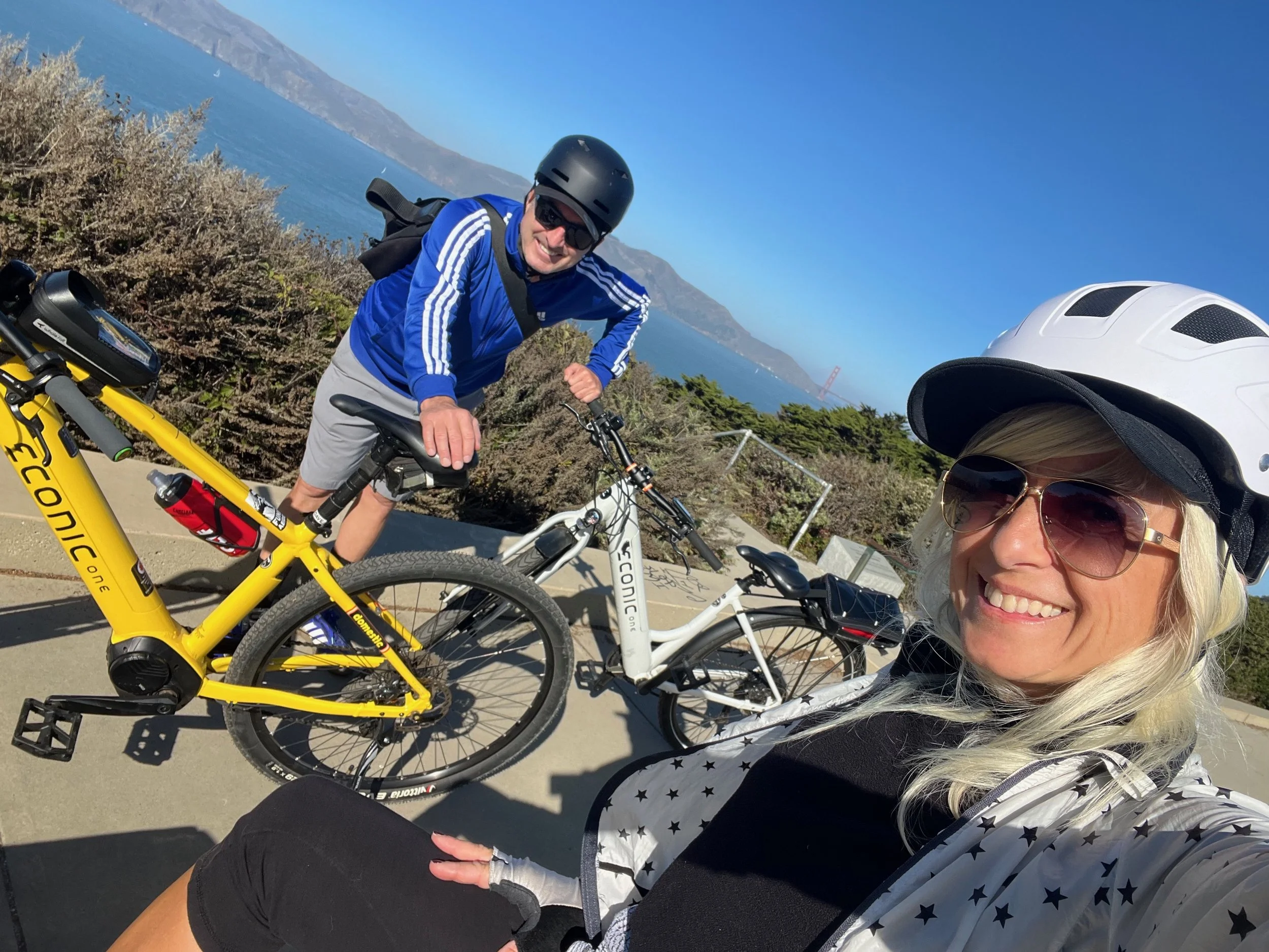 A smiling woman wearing a white helmet and sunglasses taking a selfie with a man in a black helmet, blue jacket, and sunglasses, both with electric bikes at a scenic overlook with water and mountains in the background.