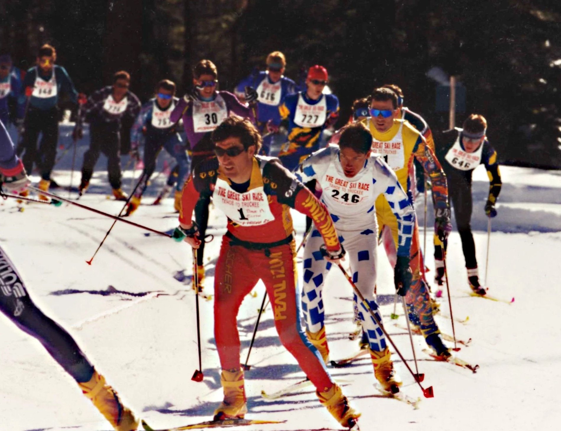 Group of people competing in a cross-country ski race, wearing colorful sports gear and numbered bibs, skiing on snow with trees in the background.
