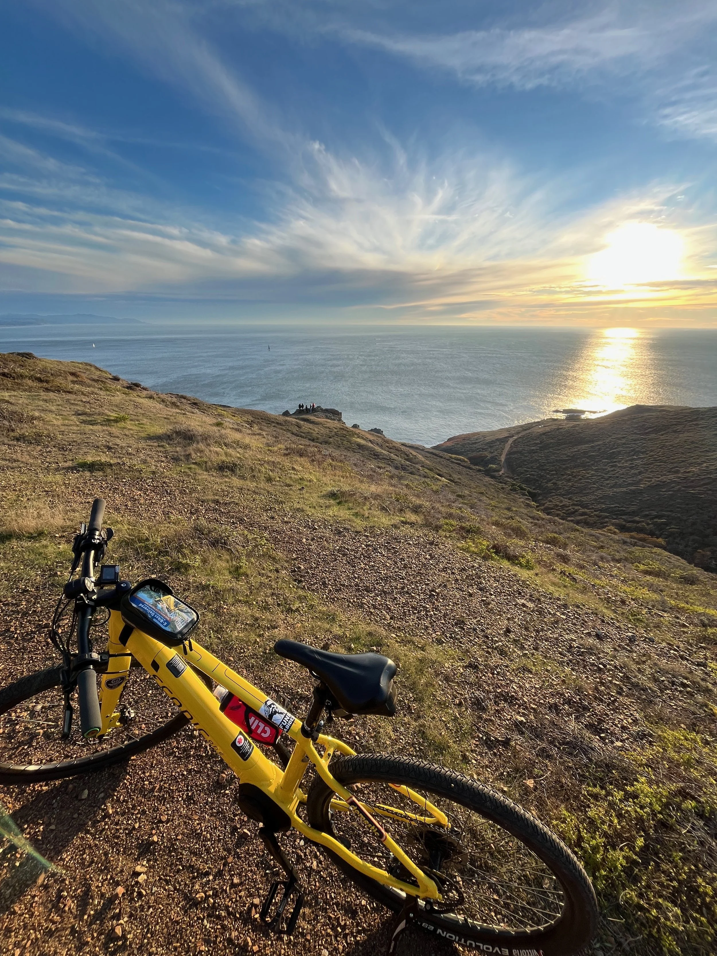 Yellow mountain bike on a trail overlooking the ocean during sunset with a partly cloudy sky.