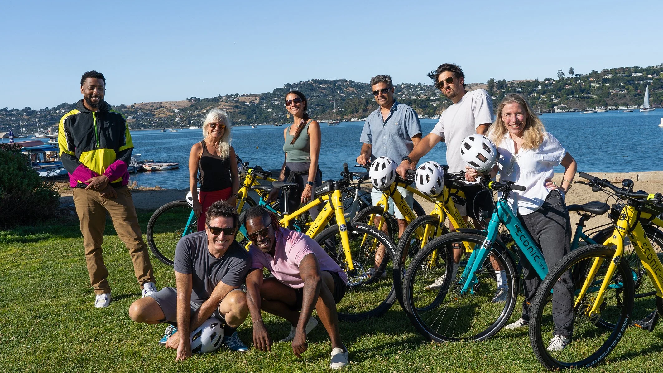 A group of nine diverse people with bicycles standing and kneeling on grass near a body of water with boats and hills in the background.