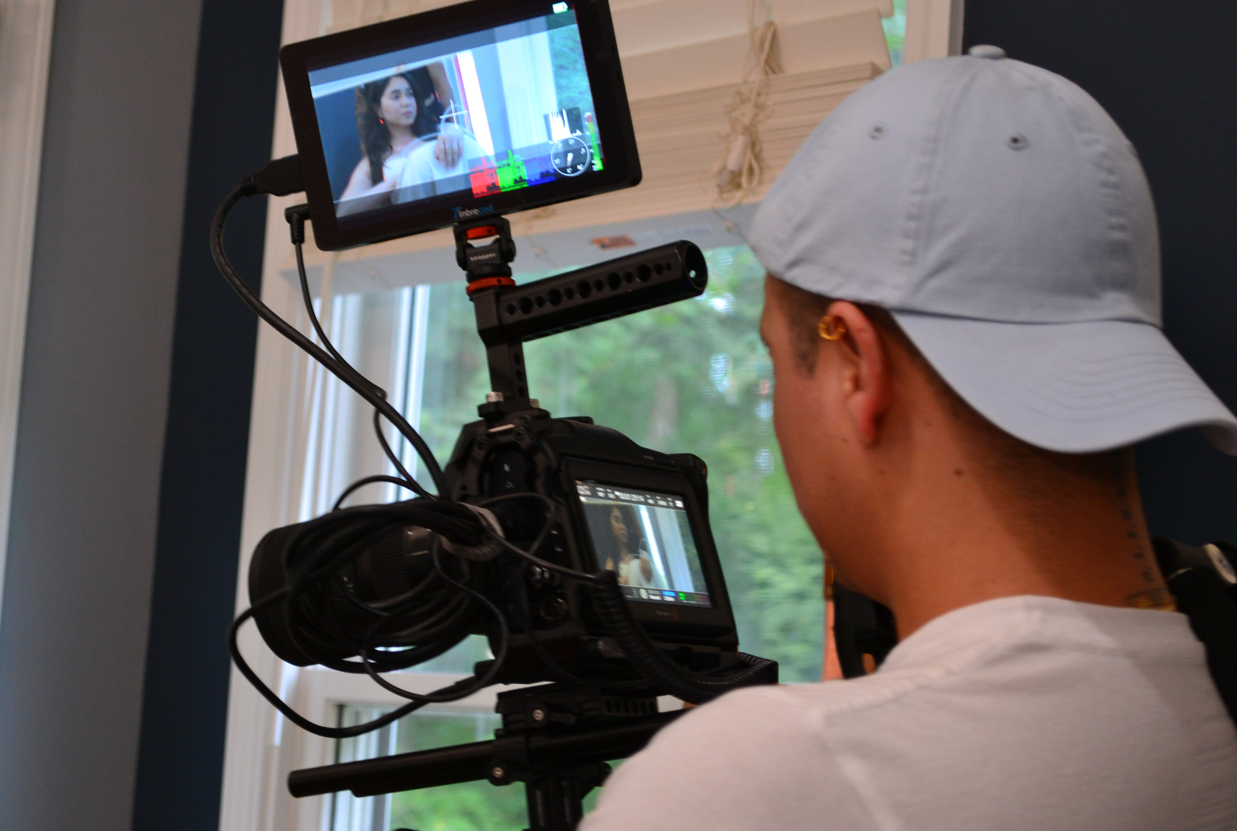 A person wearing a light-colored baseball cap and white shirt operating a professional video camera with an external monitor, filming a woman sitting by a window.