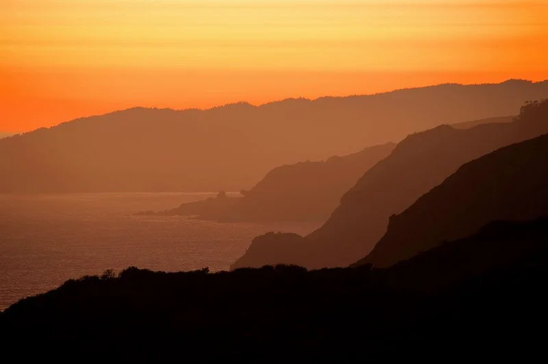 “A breathtaking view of the Marin Headlands at sunset, with layers of rugged coastal hills fading into the distance. The sky glows in shades of orange and yellow, and the Pacific Ocean lies calm beneath the golden horizon.”