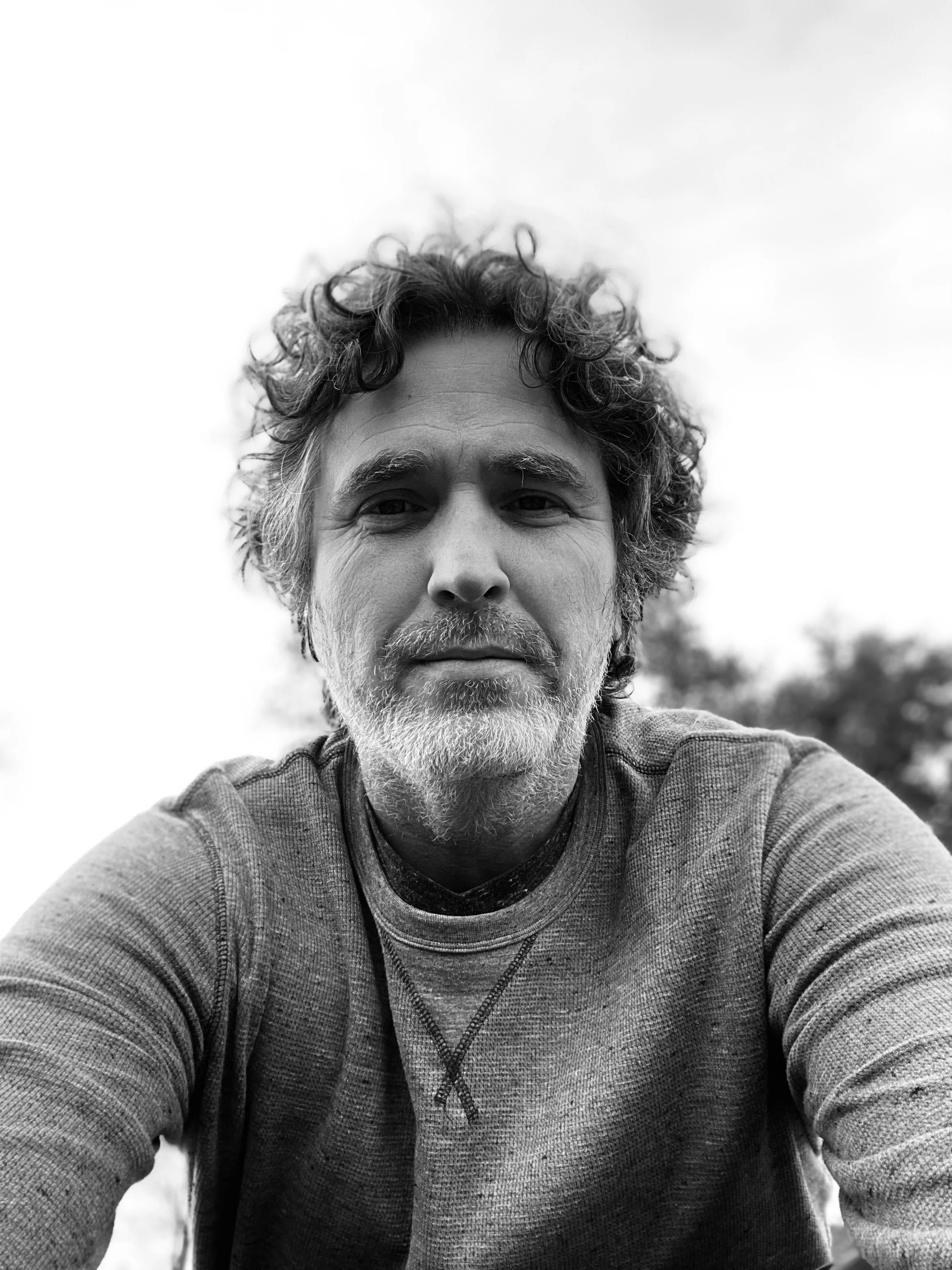 Black and white portrait of a middle-aged man with curly hair and a beard, looking at the camera with a neutral expression. He is outdoors with a blurred background of trees and a cloudy sky.