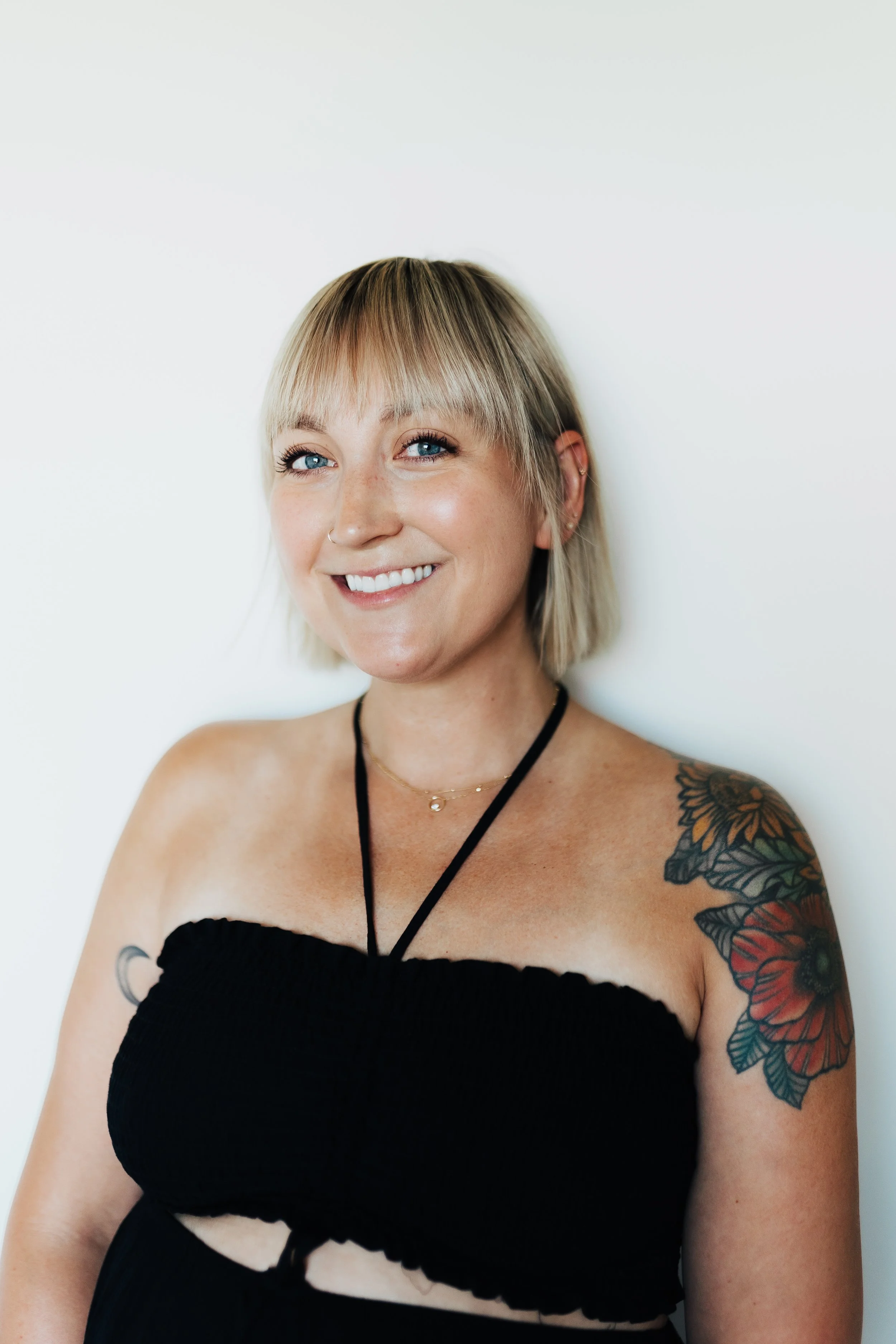 A smiling woman with short blonde hair and tattoos on her right arm, wearing a black strapless top and a delicate necklace, standing against a plain white background.