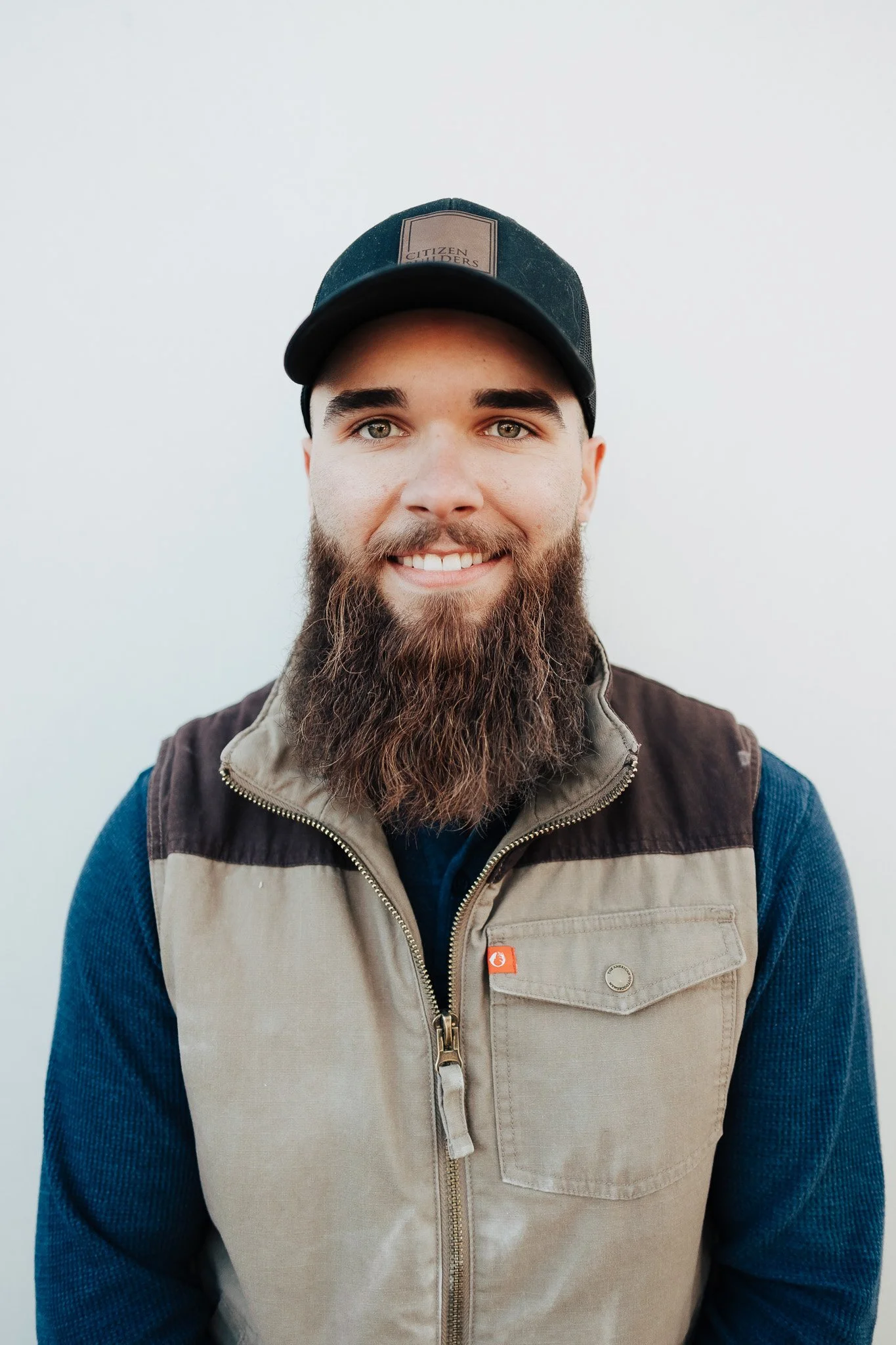 Young man with a beard smiling, wearing a black baseball cap and a beige and dark brown vest over a blue long-sleeve shirt, standing against a plain white background.