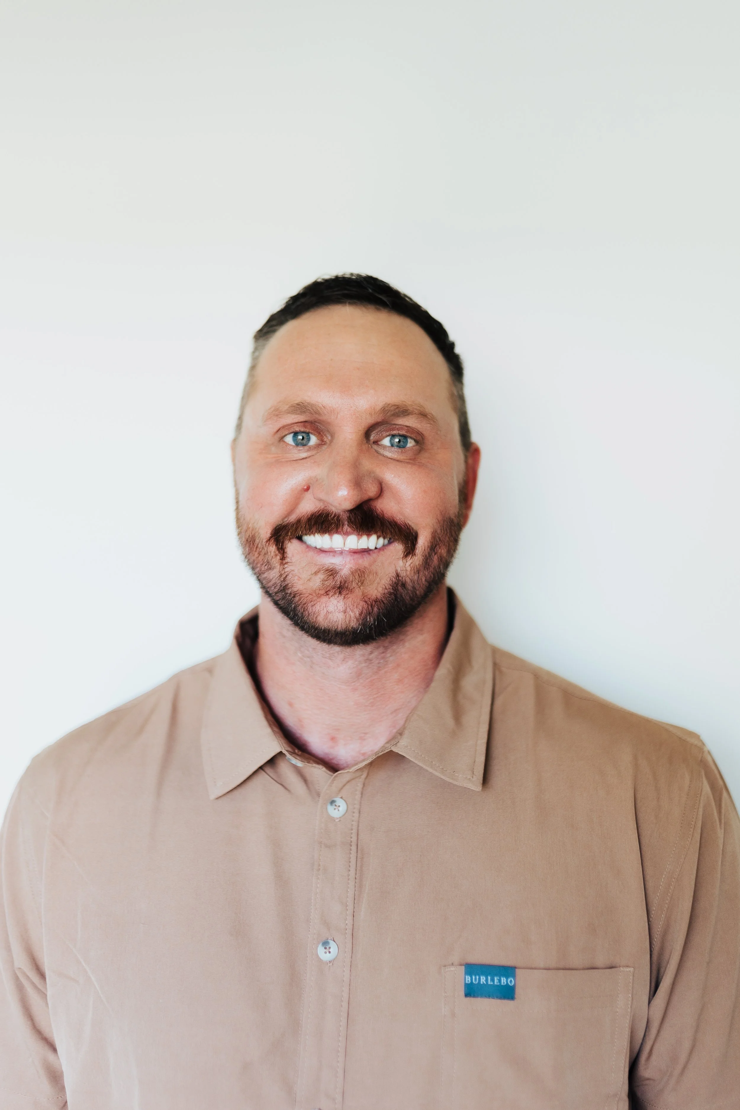 A smiling man with blue eyes, a beard, and short dark hair, wearing a beige collared shirt, standing against a plain white wall.