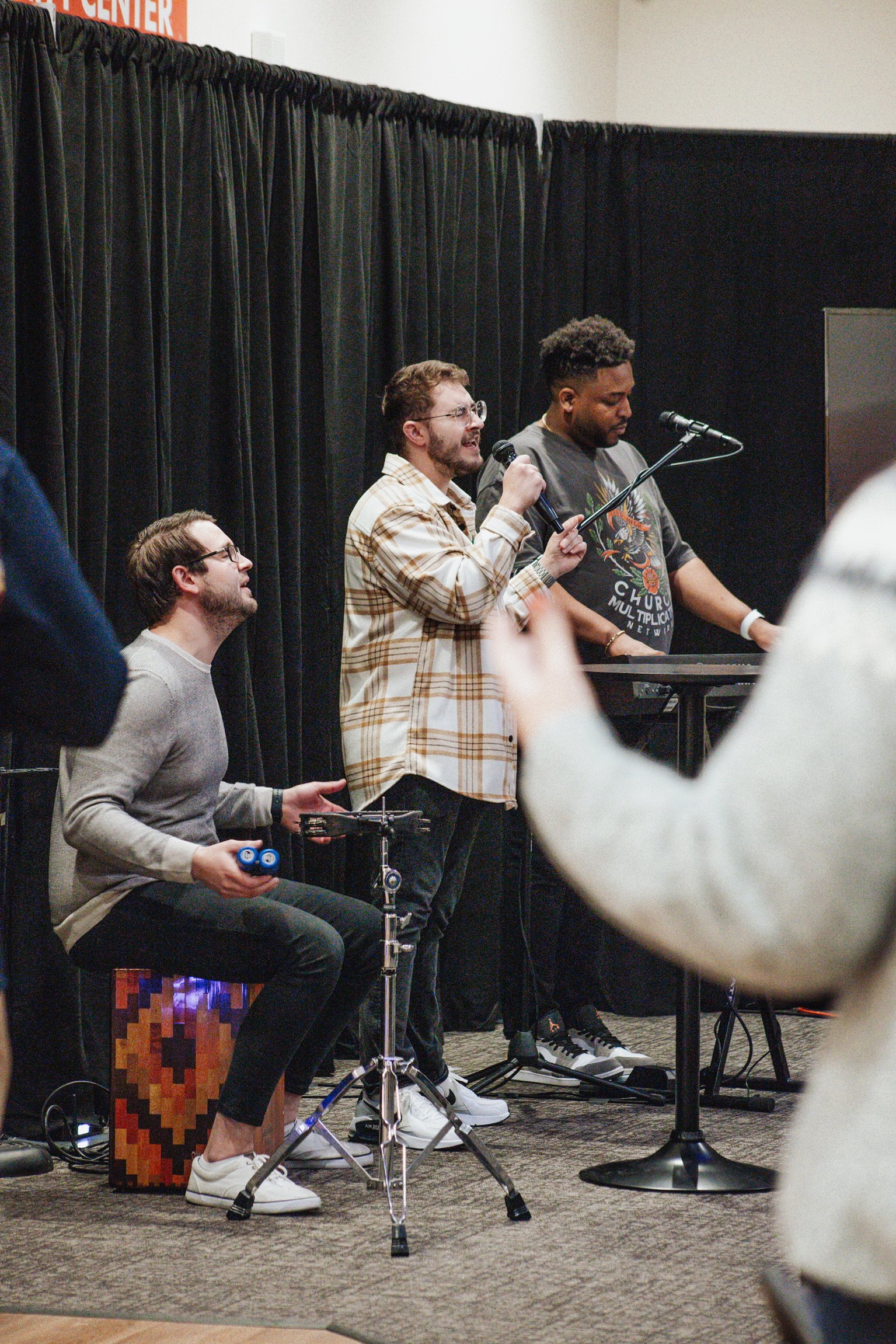 Three men performing on stage with black curtains in the background. One man is playing a percussion instrument, another is singing into a microphone, and the third is playing a keyboard. An audience member's arm is visible in the foreground.