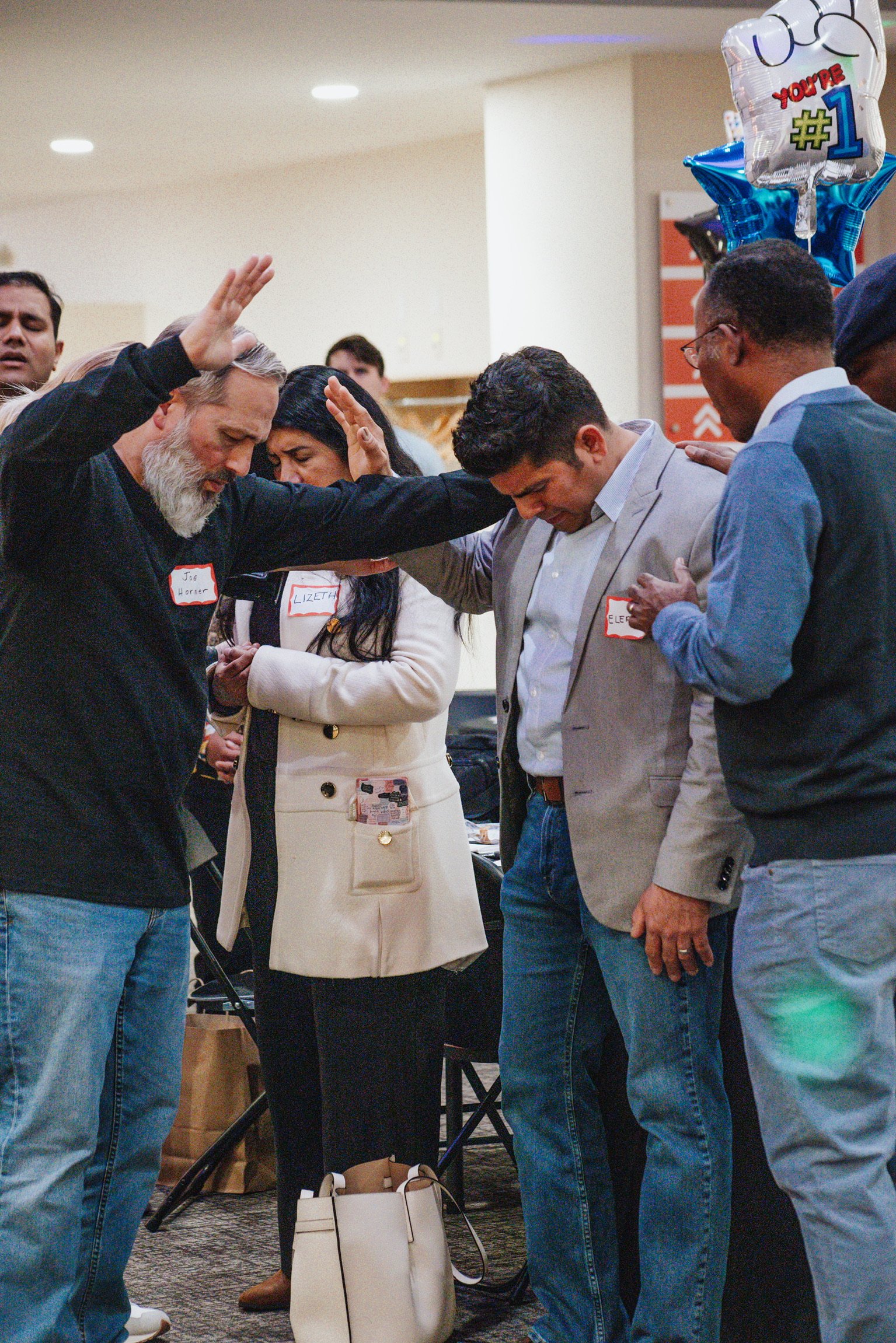 Group of people praying with heads bowed, some with hands raised, in a casual indoor setting.