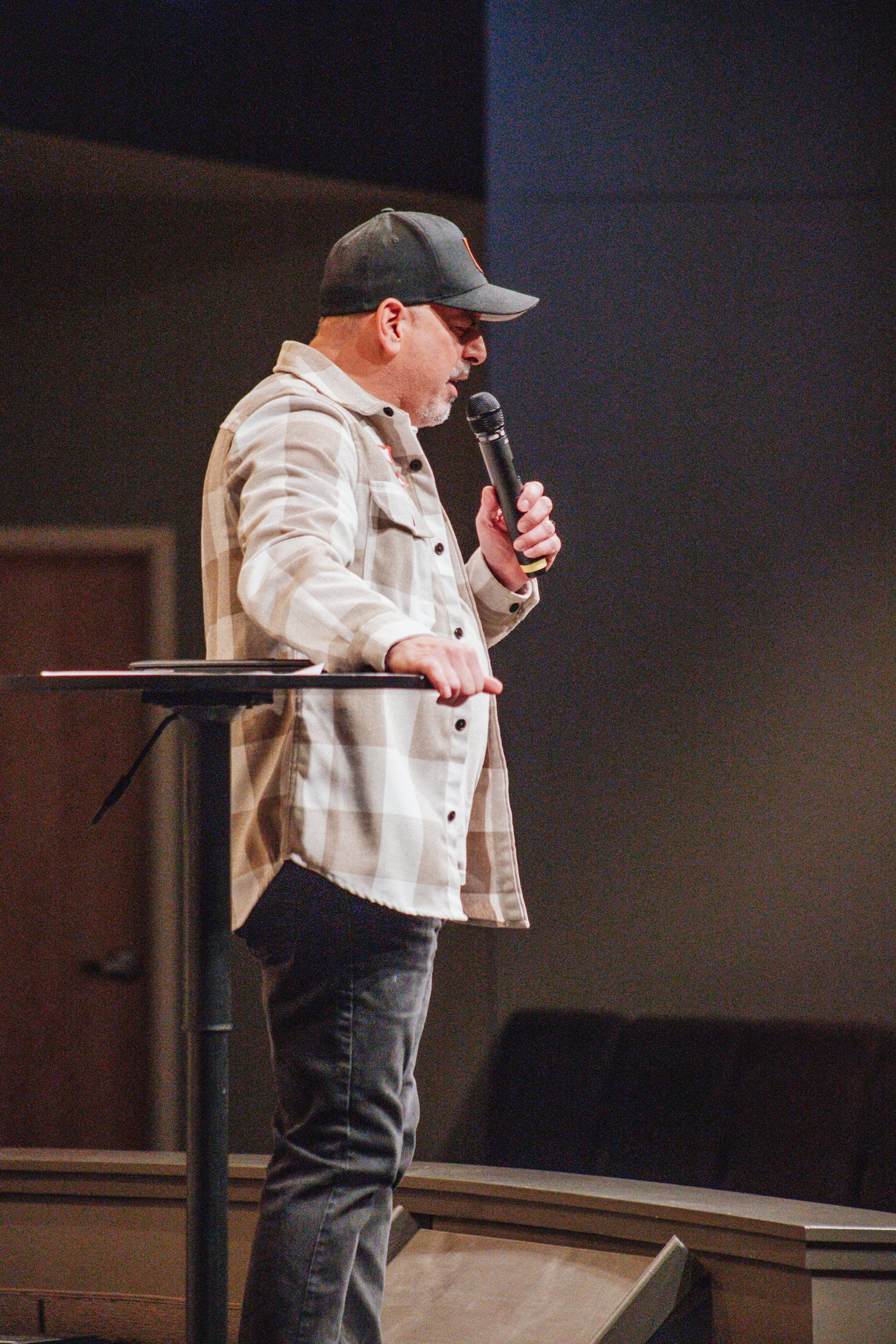 A man wearing a black cap, glasses, and a beige plaid shirt holding a microphone and standing beside a podium in a dimly lit room.