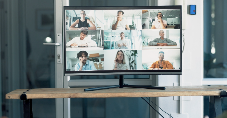 A computer monitor on a wooden desk displaying a video conference with nine diverse participants in a grid layout, each in different indoor settings.