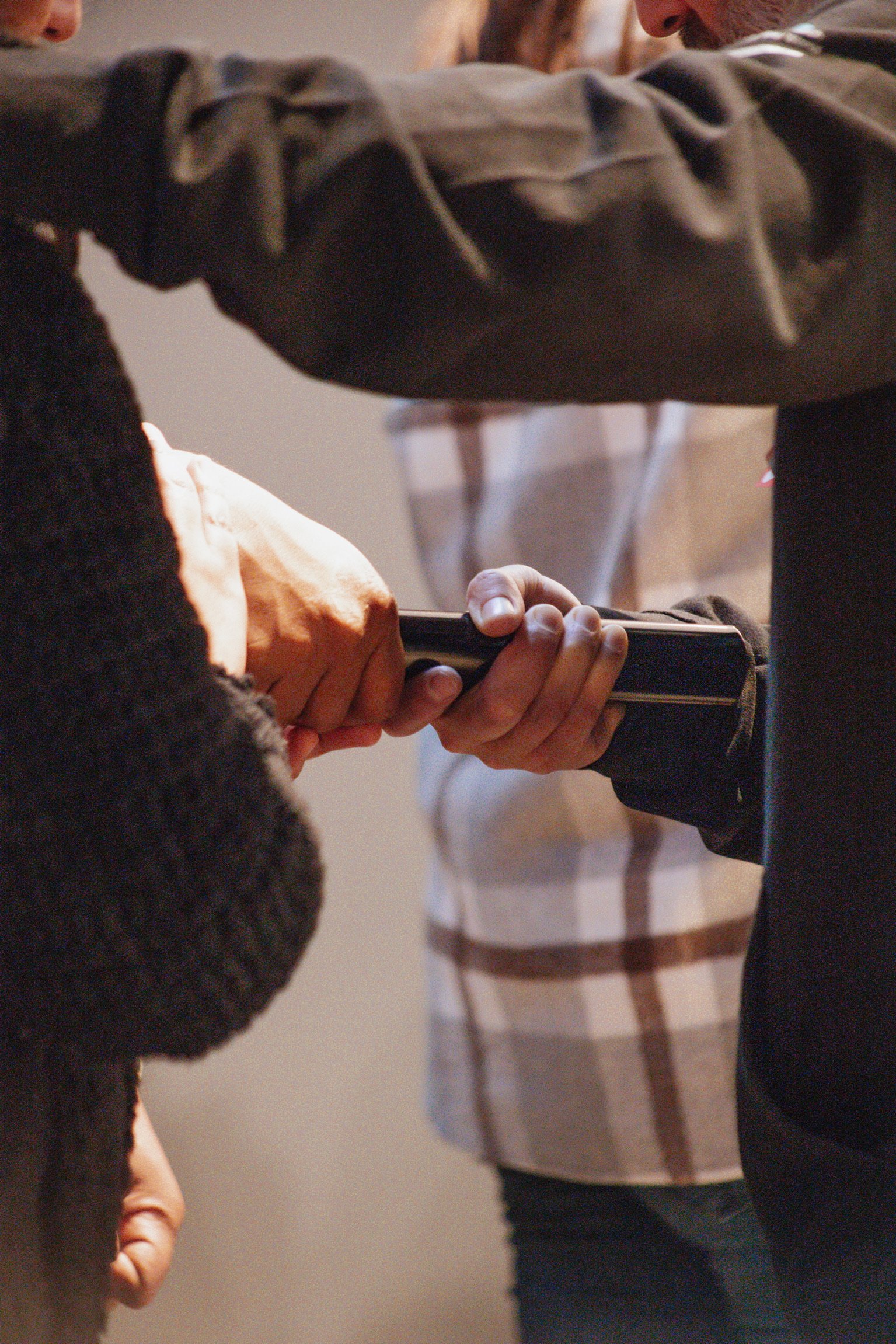 A person is handing over a black wallet to another person with a plaid shirt, with a child in a black sweater observing. Only parts of their bodies are visible.