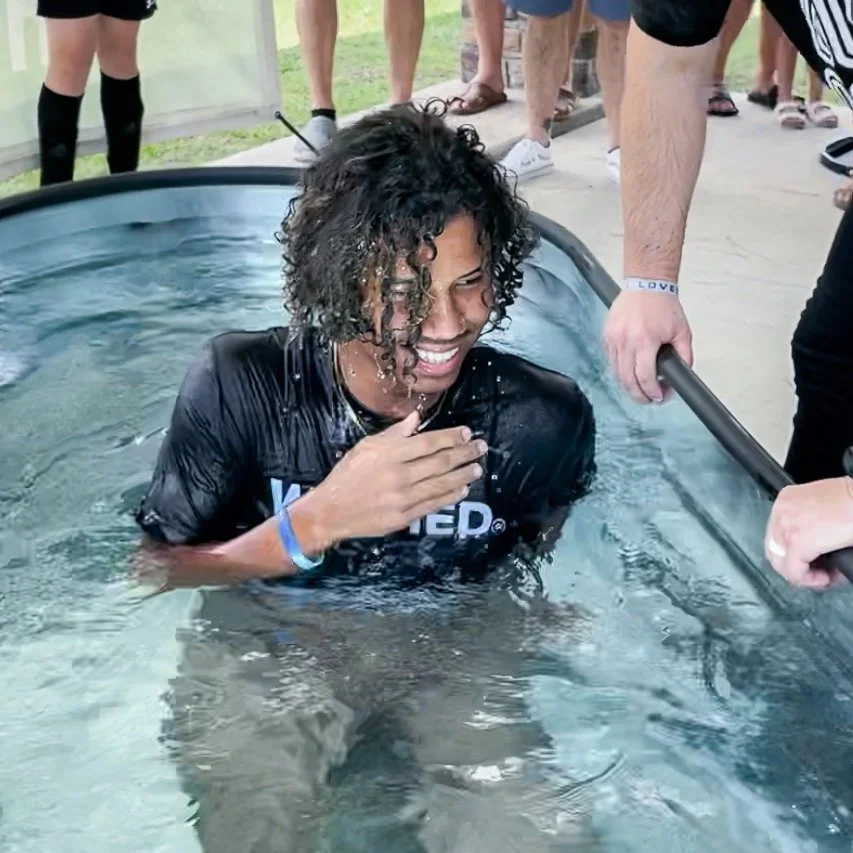 Person standing smiling in a large tub of water, surrounded by others, likely during a baptism or water event.