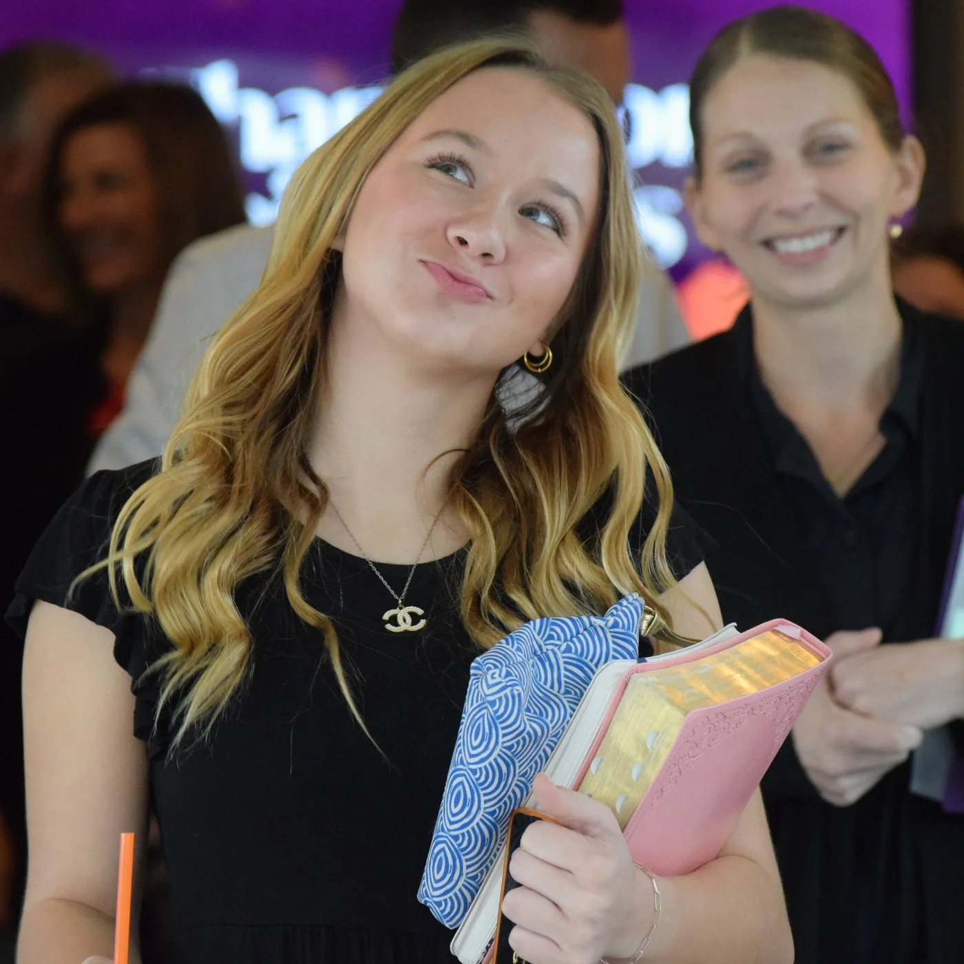 Young woman with long hair holding books and making a playful expression, with a person smiling in the background.