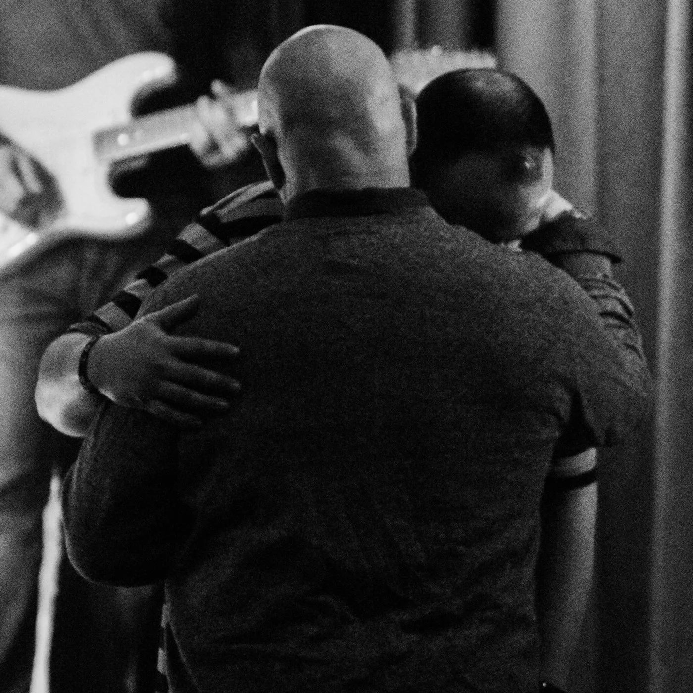 Two people hugging closely, with the focus on their backs, in a dimly lit environment with a guitar player in the background.