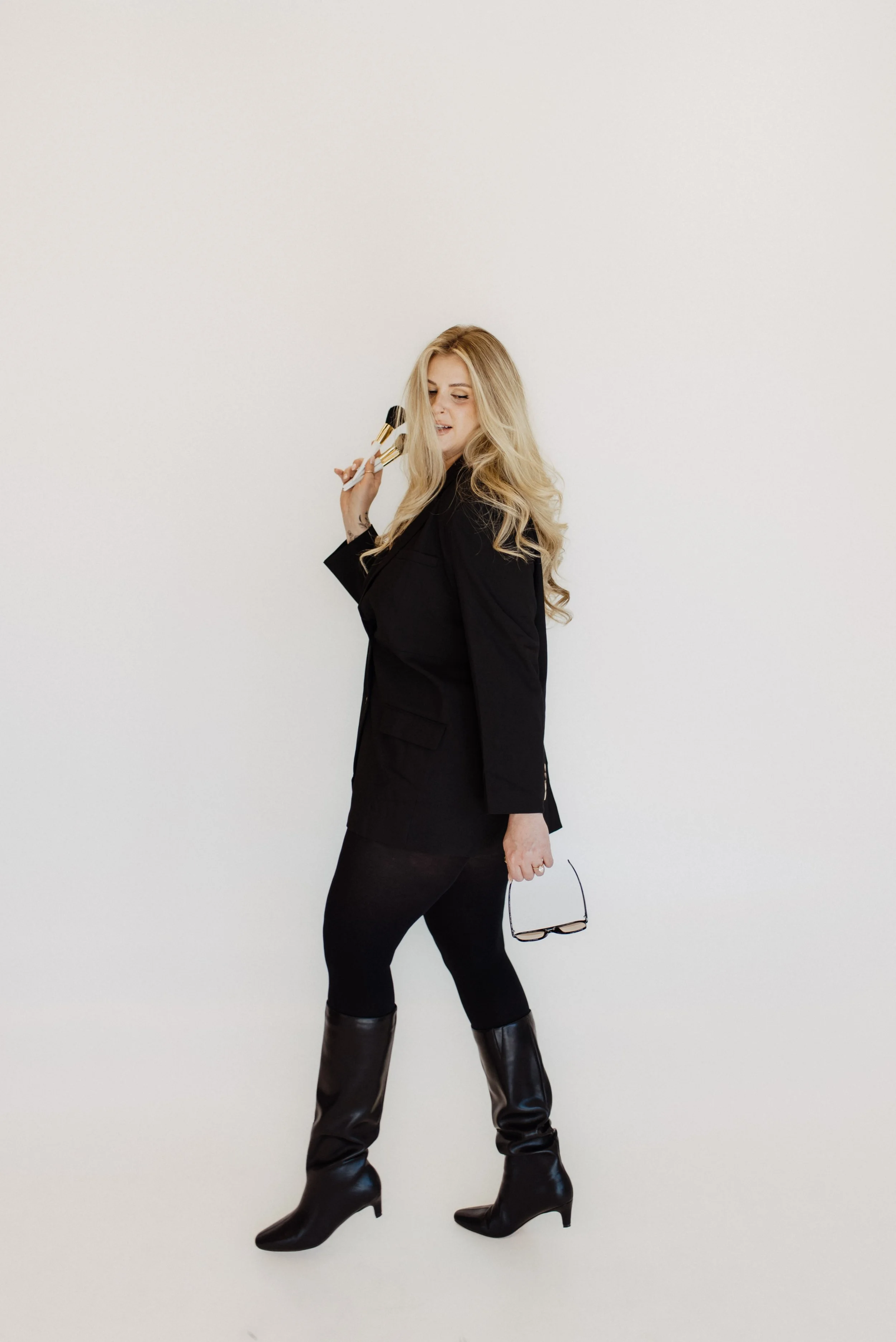 A woman with long blonde hair wearing a black blazer, black tights, and black high-heeled boots, standing against a plain white background, holding a pair of glasses in her left hand and a makeup brush in her right hand.