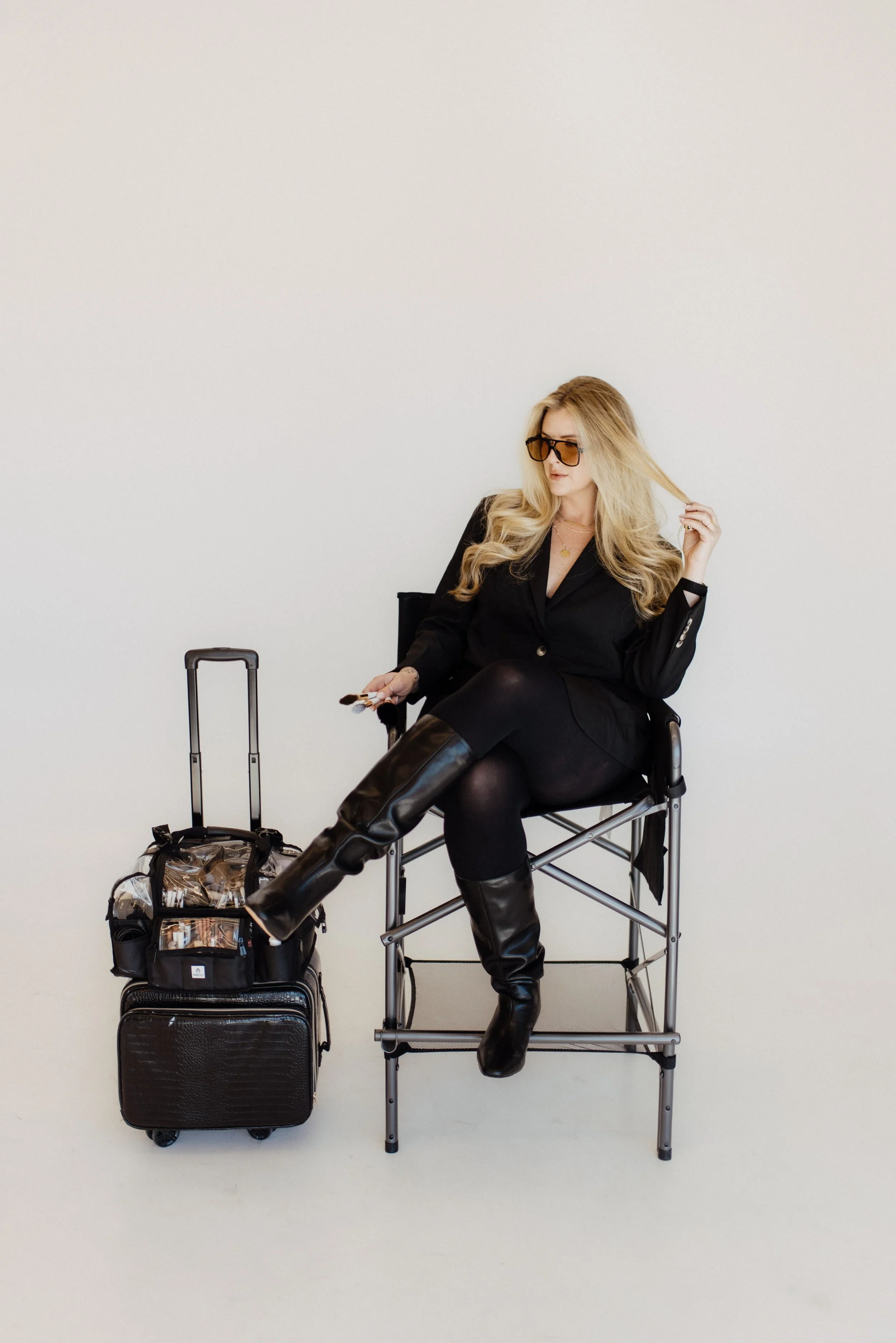 A woman with long blonde hair wearing black sunglasses, a black blazer, black leggings, and black knee-high boots sitting on a folding chair in front of a plain white background. She has a small suitcase and a rolling carry-on bag beside her.