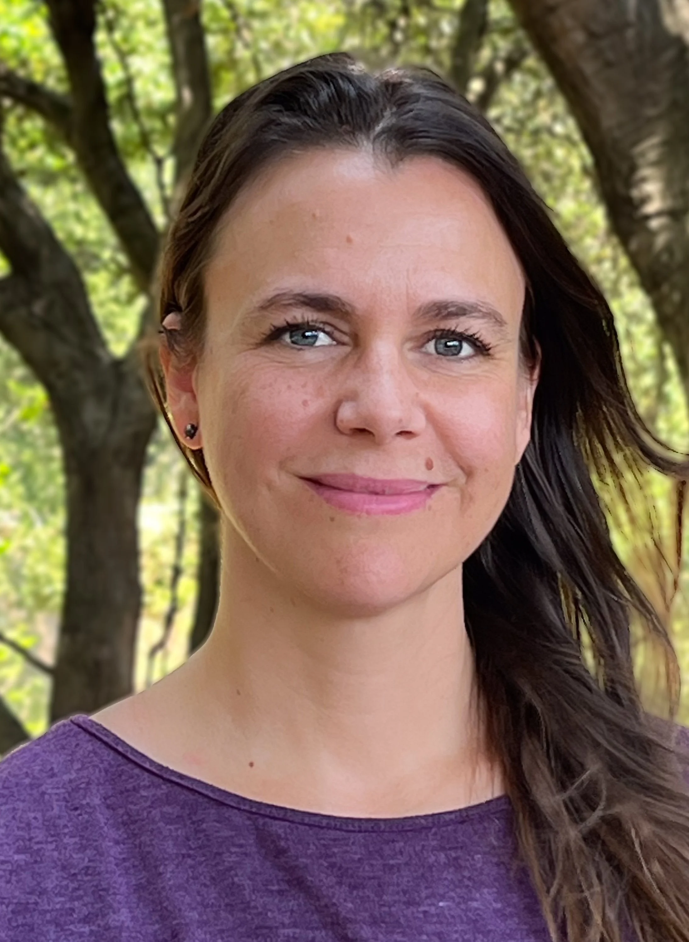 A woman with dark hair, blue eyes, and small earrings outside in front of green trees, smiling at the camera.