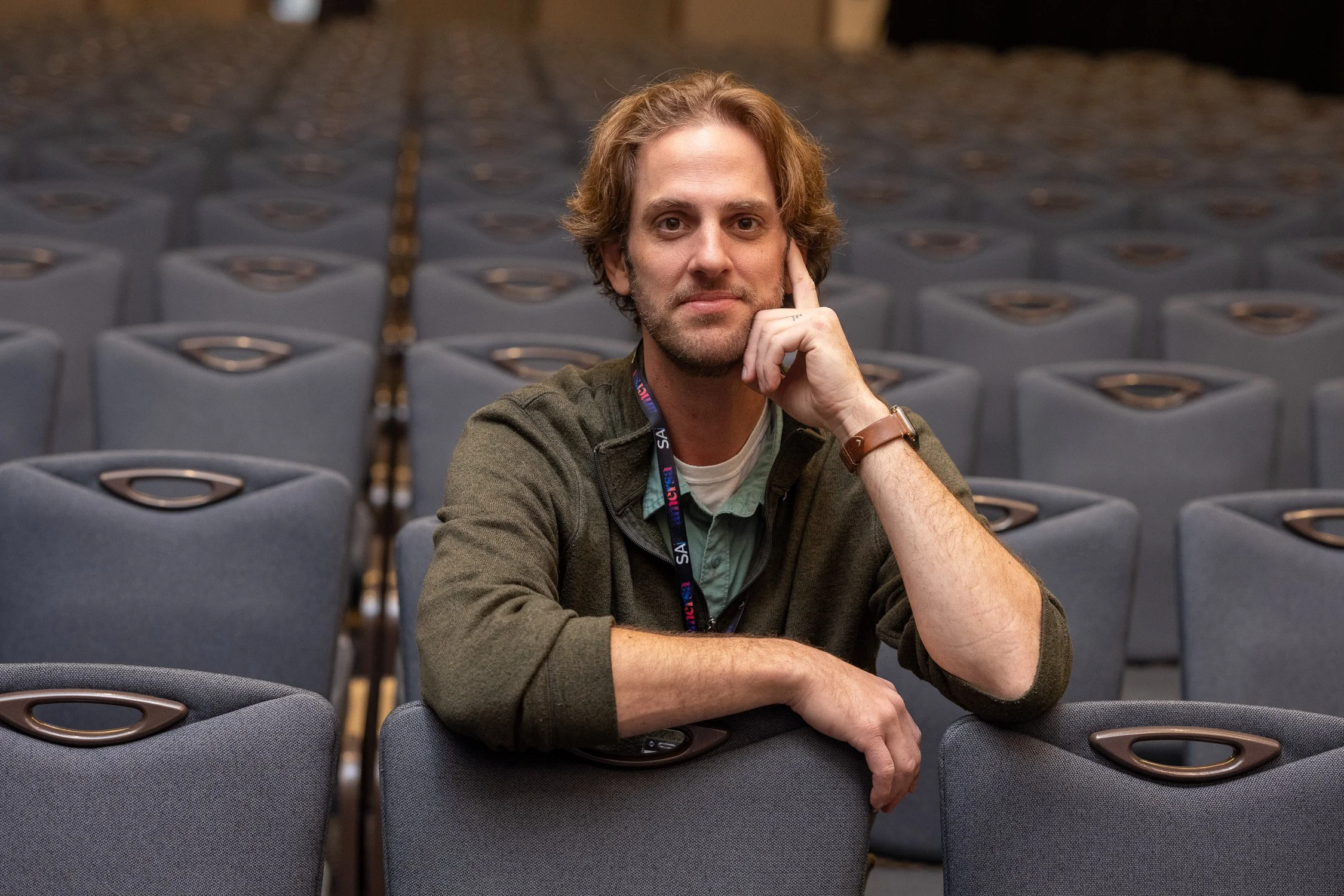 A man with curly hair, a beard, and a casual green jacket sitting alone in an empty auditorium or conference hall with rows of gray chairs behind him.